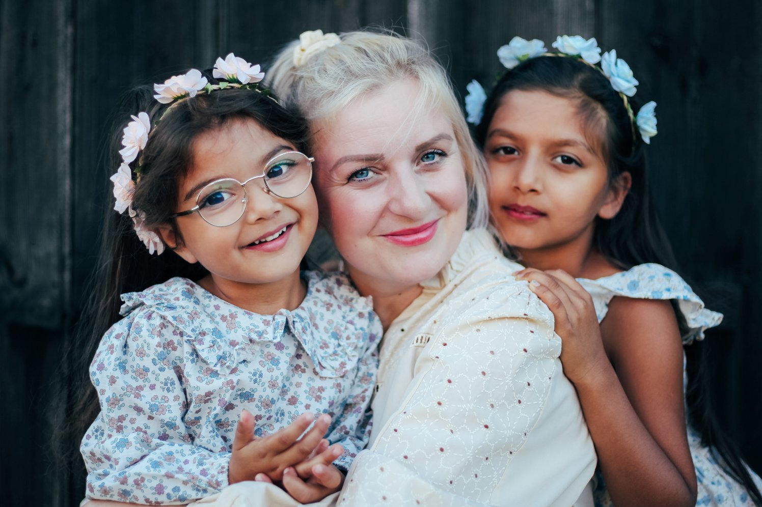 Portret matki z dwiema córkami w kwiecistych wiankach na tle ciemnego drewna. Dziewczynka w okularach uśmiecha się szeroko.