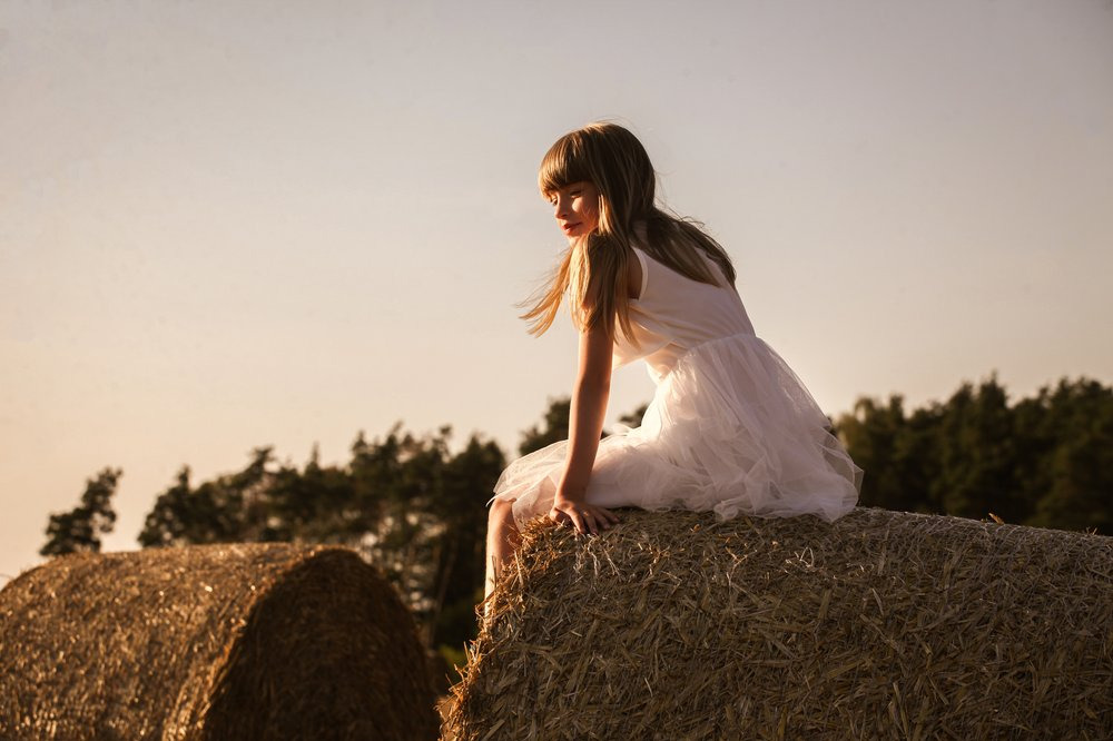 Dziewczynka w białej sukience siedzi na słomianej beli na tle pola i lasu w ciepłym świetle zachodzącego słońca. Delikatny portret w plenerze.