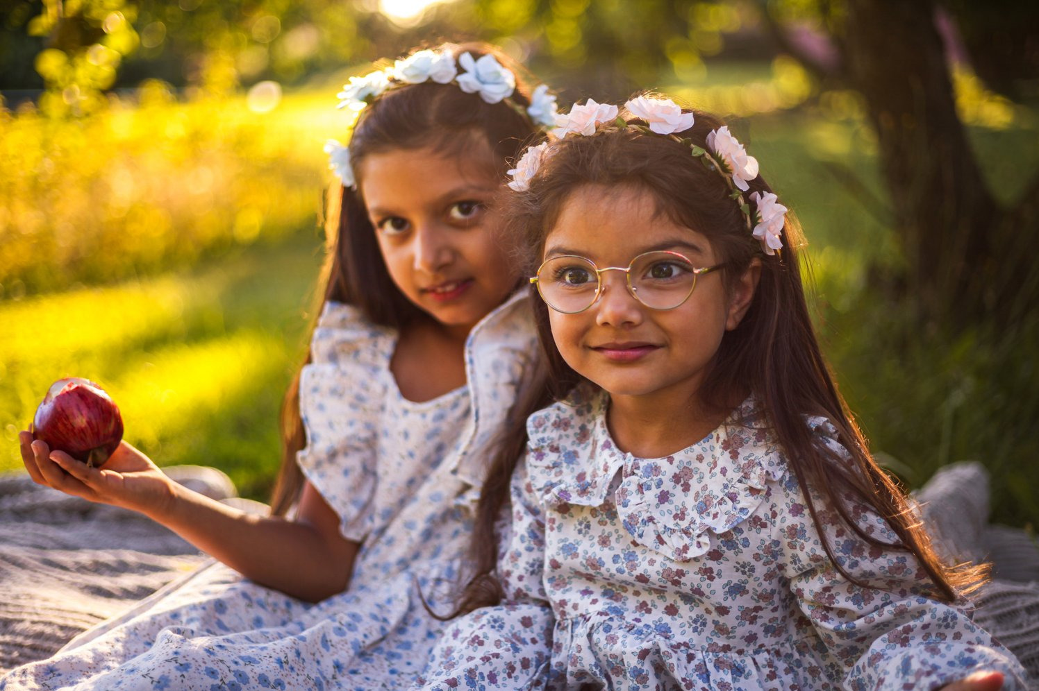 Dwie urocze dziewczynki w kwiecistych sukienkach i wiankach na głowach, siedzą na kocu w sadzie, jedna trzyma jabłko. Ciepłe, naturalne światło.