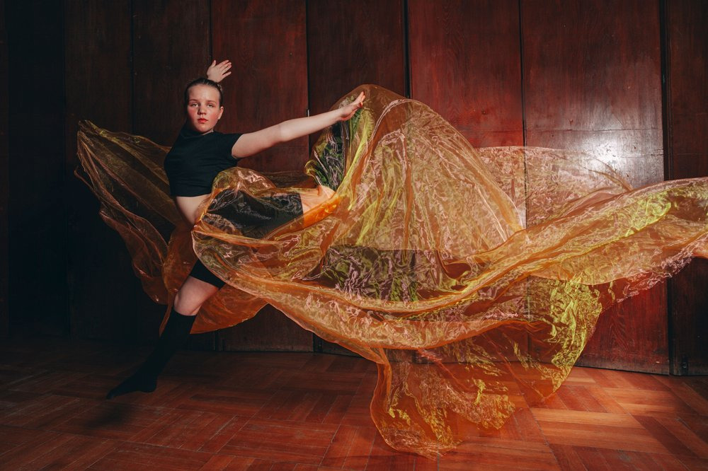 Dynamiczne ujęcie kobiety w tańcu z pomarańczową, zwiewną tkaniną na tle drewnianej ściany i parkietu. Ekspresja ruchu, artystyczna fotografia.