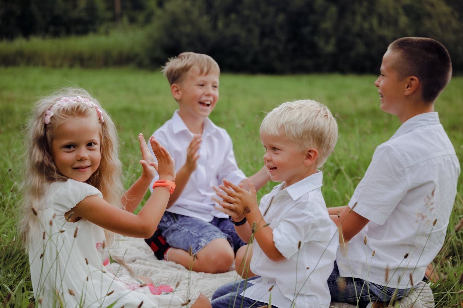 Uśmiechnięta dziewczynka z opaską i chłopcy w białych koszulach siedzą na kocu w trawie, klaszcząc w dłonie. Radosna atmosfera rodzinnego spotkania na łonie natury.