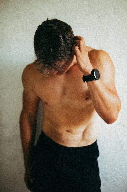 Mężczyzna bez koszulki, w czarnych spodniach, z zegarkiem na ręce, dotyka włosów na tle jasnej ściany. Ujęcie z góry, miękkie światło, widoczne niedoskonałości skóry.