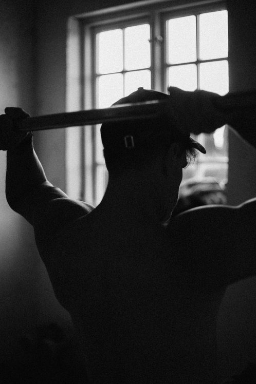 Czarno-białe zdjęcie sylwetki mężczyzny ćwiczącego na drążku w Rumi, na tle okna z kratami, ukazujące jego muskulaturę.