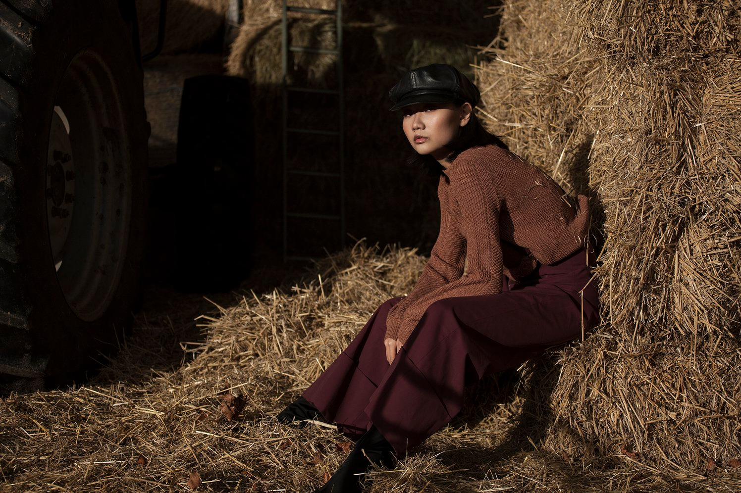 Stylowa sesja: Azjatka w brązowym swetrze i czarnej czapce siedzi na sianie przy oponie traktora. Ciemne, nastrojowe oświetlenie podkreśla rustykalny charakter.