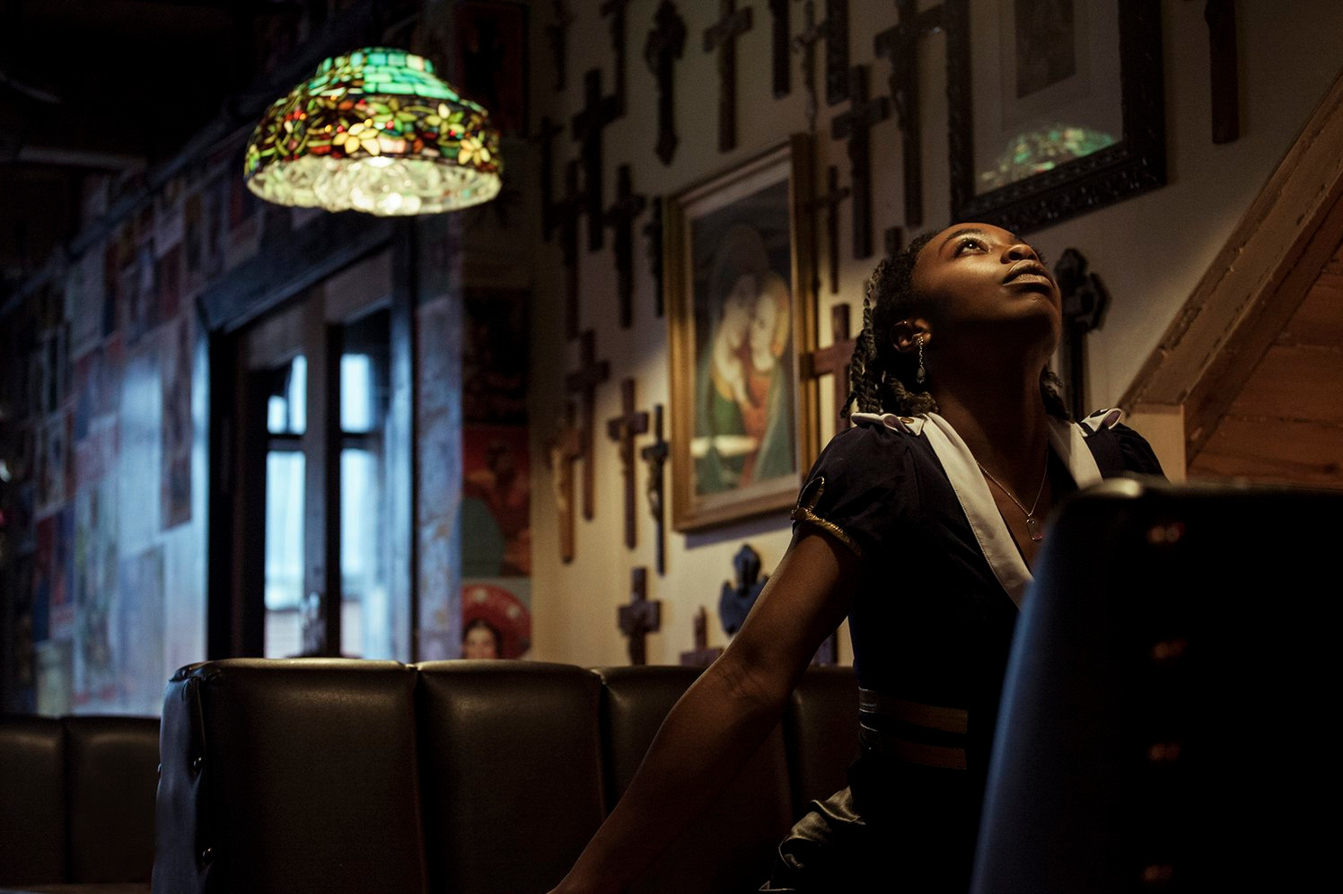 Portret kobiety spoglądającej w górę, w tle ściana z krzyżami i witrażowa lampa, tworzące unikalny klimat wnętrza. Ujęcie z dołu podkreśla ekspresję modelki.