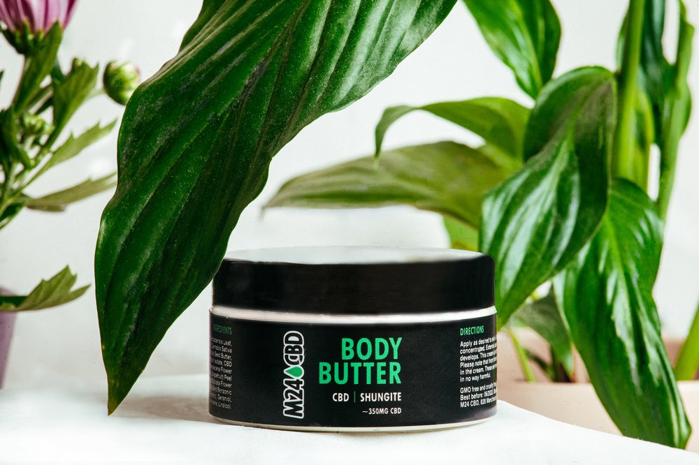 Słoik z masłem do ciała CBD na białym tle, otoczony zielonymi liśćmi roślin. Etykieta z napisem 'Body Butter' i informacjami o składnikach. Stonowana kolorystyka, naturalne światło.