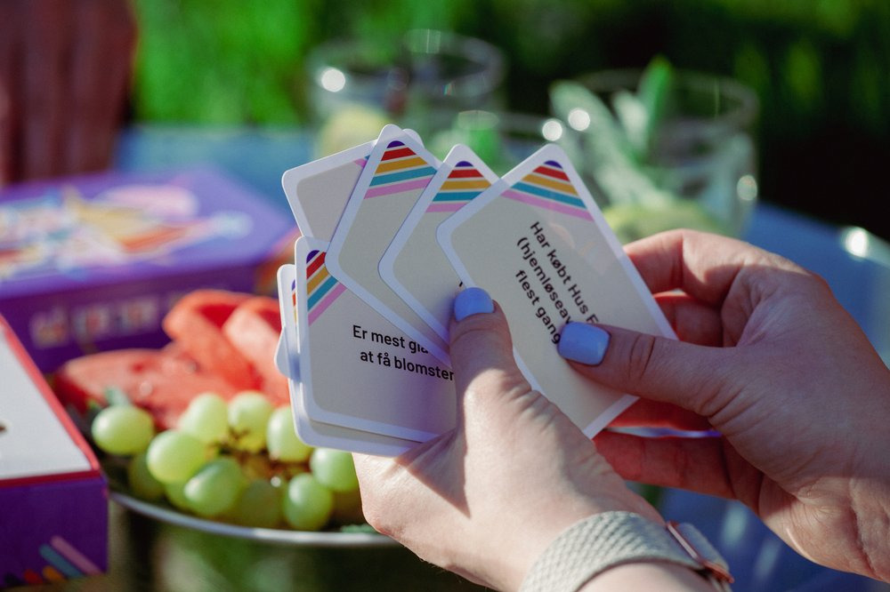 Dłoń trzymająca karty do gry z kolorowymi paskami na tle owoców i pudełka gry, ukazująca chwile relaksu i zabawy na świeżym powietrzu.