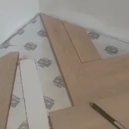 Układanie paneli podłogowych w rogu pokoju na podkładzie z nadrukiem, widoczny ołówek i fragment dociętej listwy przypodłogowej. Prace w toku.