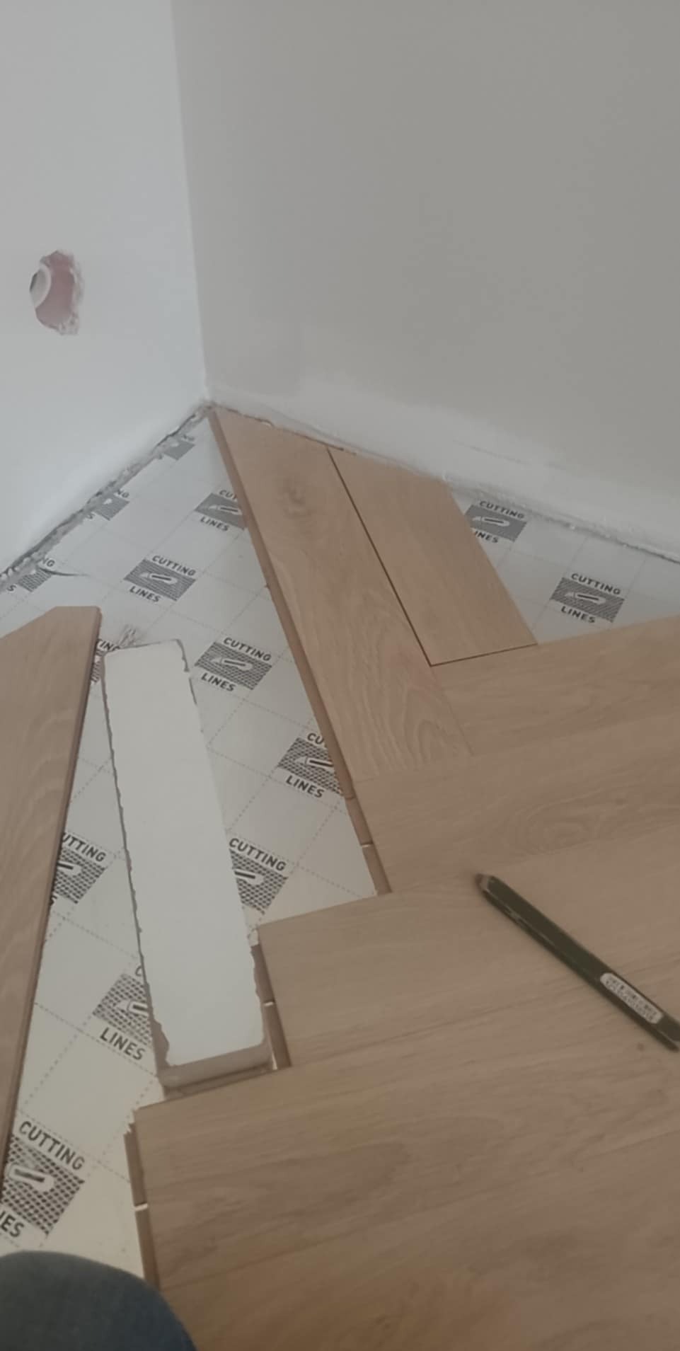 Układanie paneli podłogowych w rogu pokoju na podkładzie z nadrukiem, widoczny ołówek i fragment dociętej listwy przypodłogowej. Prace w toku.