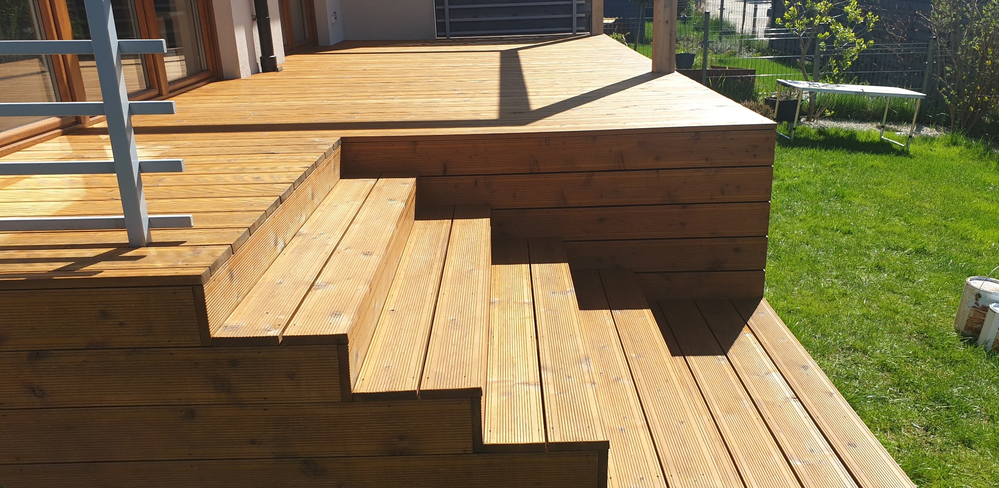 Jasnobrązowy taras drewniany z schodami, otoczony zielonym trawnikiem. Widoczne deski ułożone równolegle, tworzące harmonijną całość z elementami architektury.