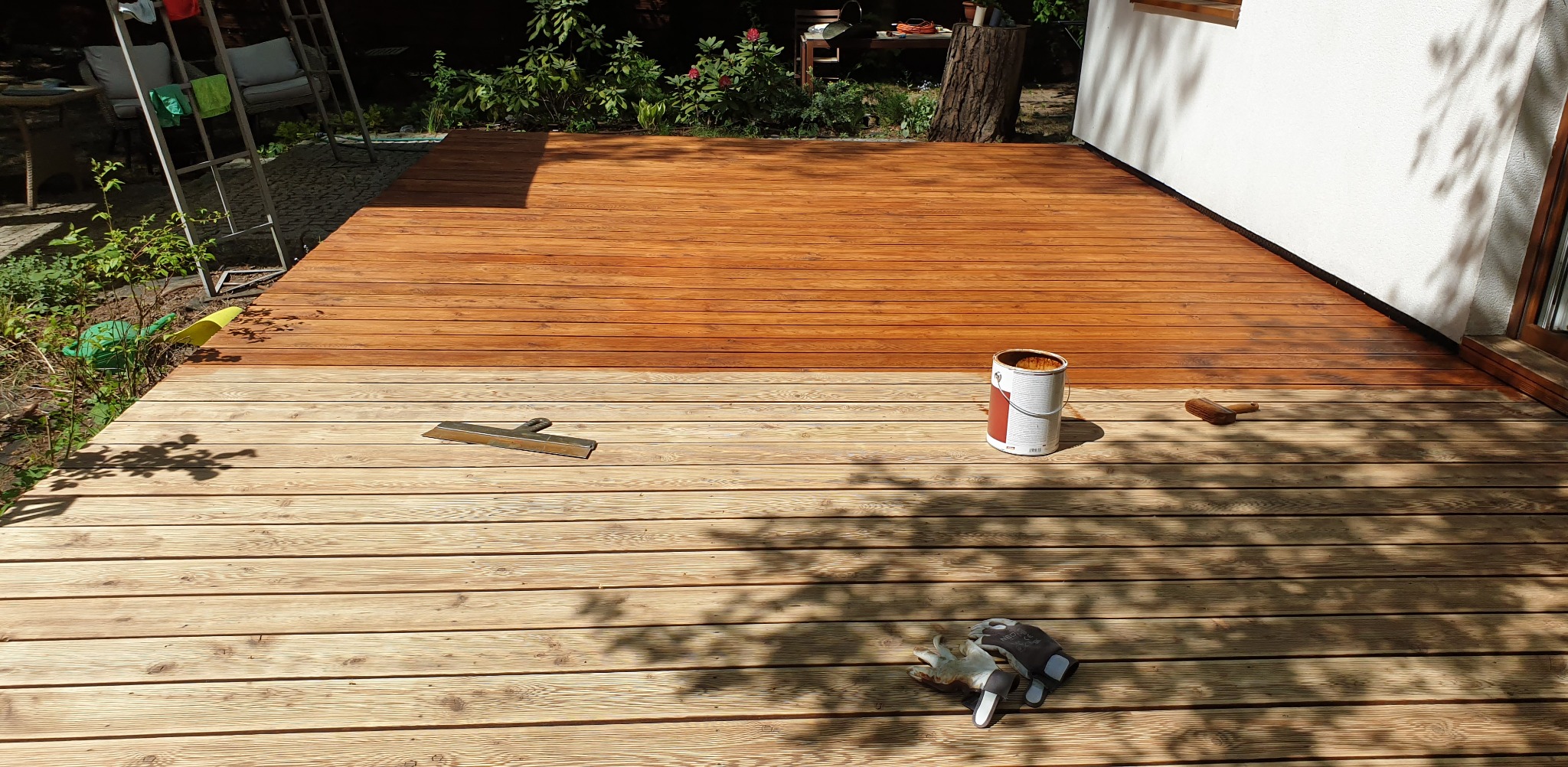 Impregnowanie drewnianego tarasu. Część desek pokryta brązowym impregnatem, obok puszka z impregnatem, pędzel i rękawice robocze. Widoczne cienie drzew na tarasie.