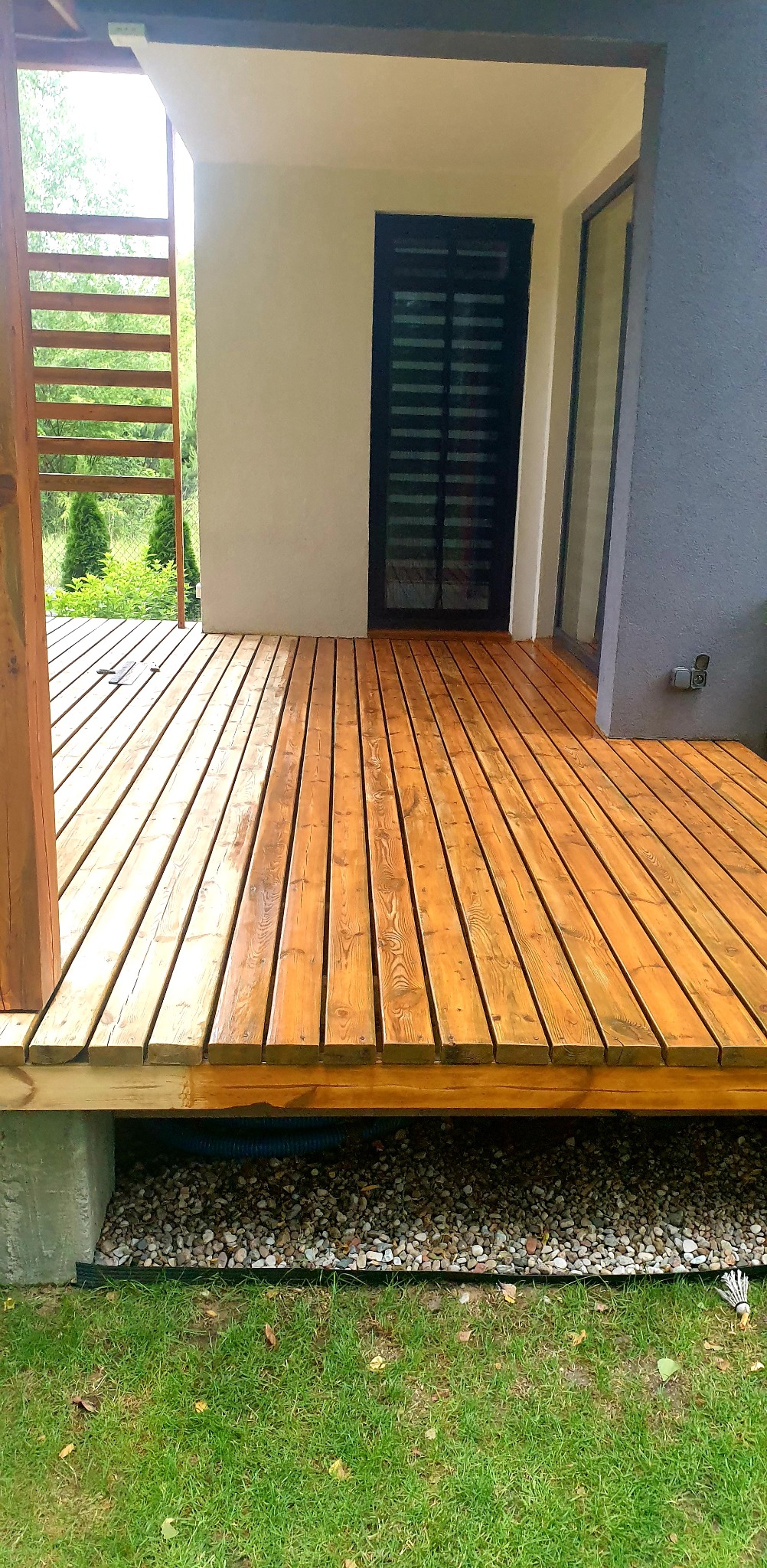 Świeżo polakierowany taras z drewna egzotycznego, z widoczną konstrukcją nośną i drenażem z kamienia. Nowoczesny budynek w tle.