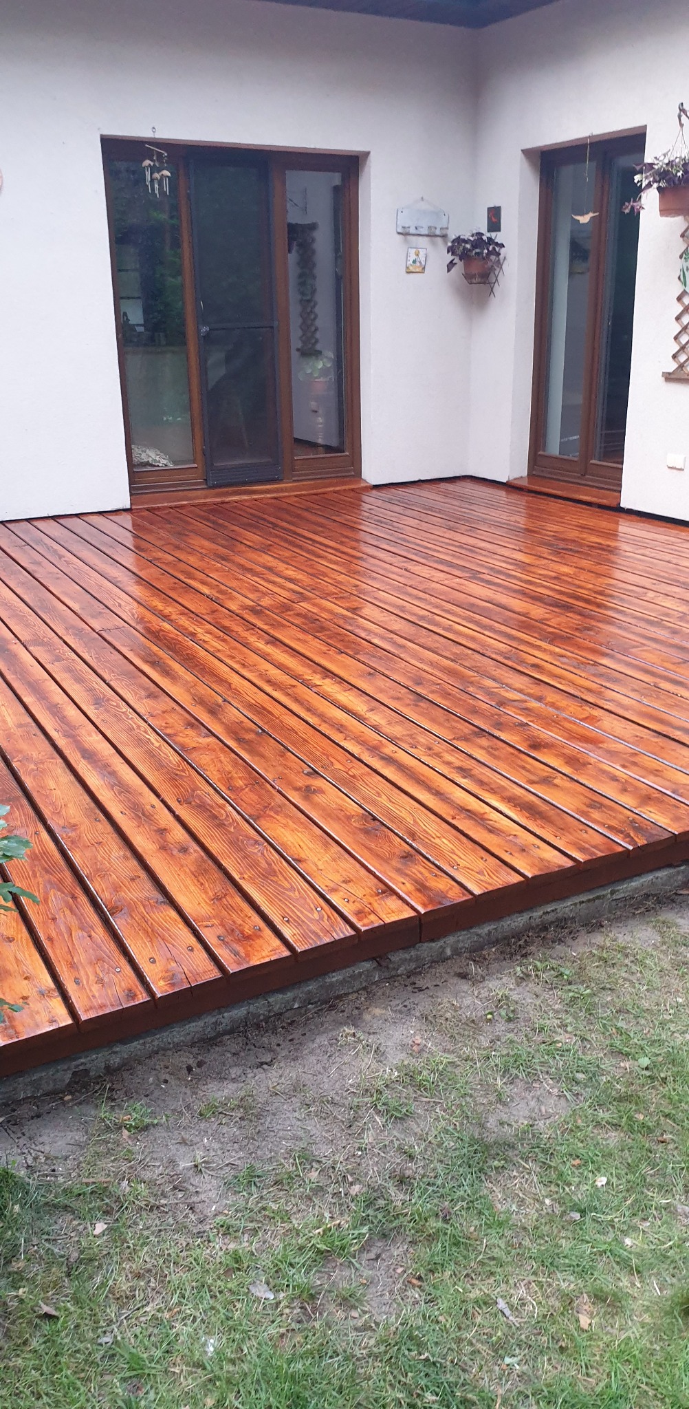 Świeżo polakierowany, drewniany taras w ciepłym odcieniu brązu, z widocznymi słojami drewna. Dwie pary drzwi balkonowych w tle, dekoracje na ścianie.