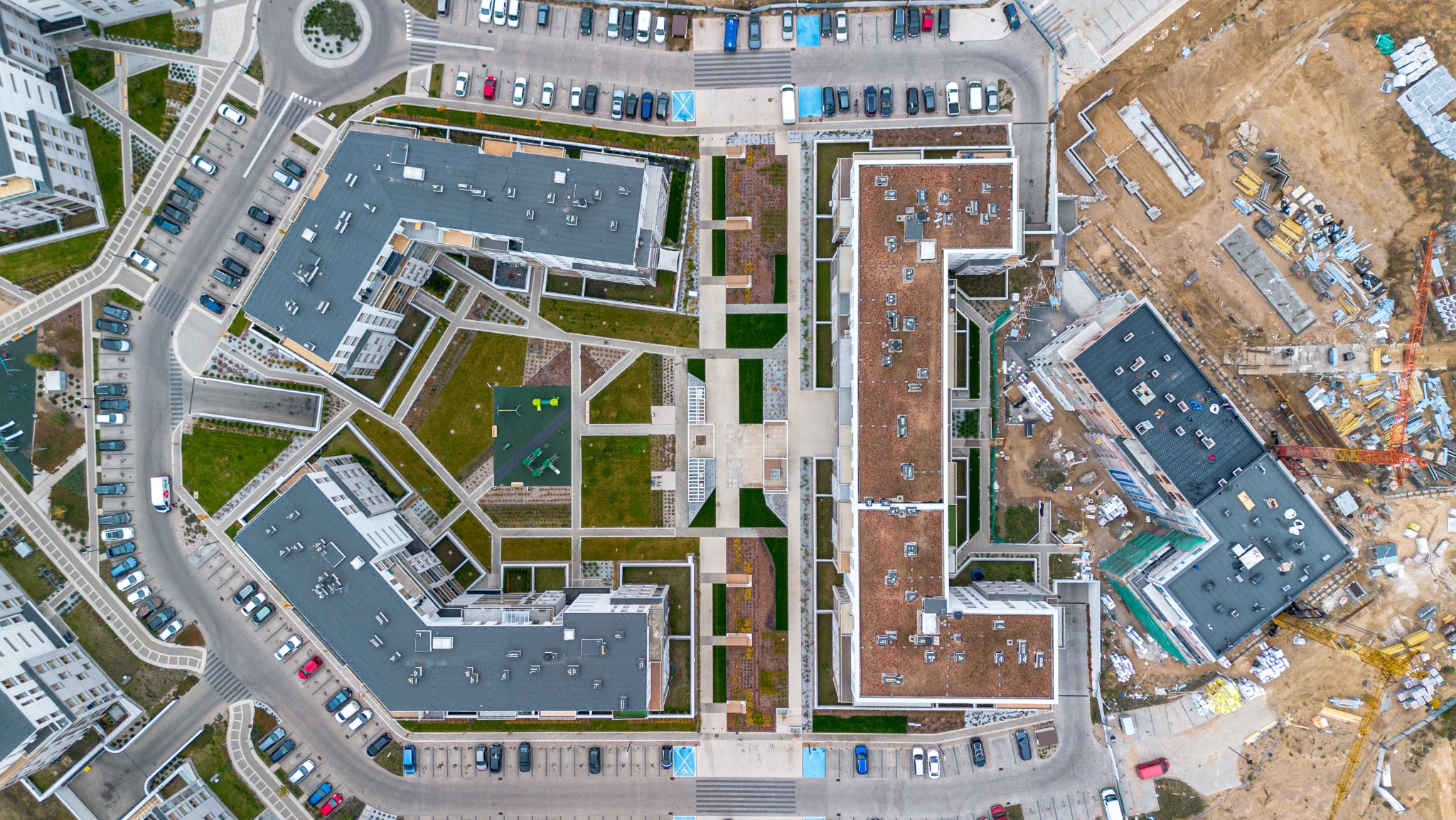 Widok z drona na osiedle mieszkaniowe z parkingami, zielenią i placem budowy w tle. Geometryczny układ budynków i przestrzeni.