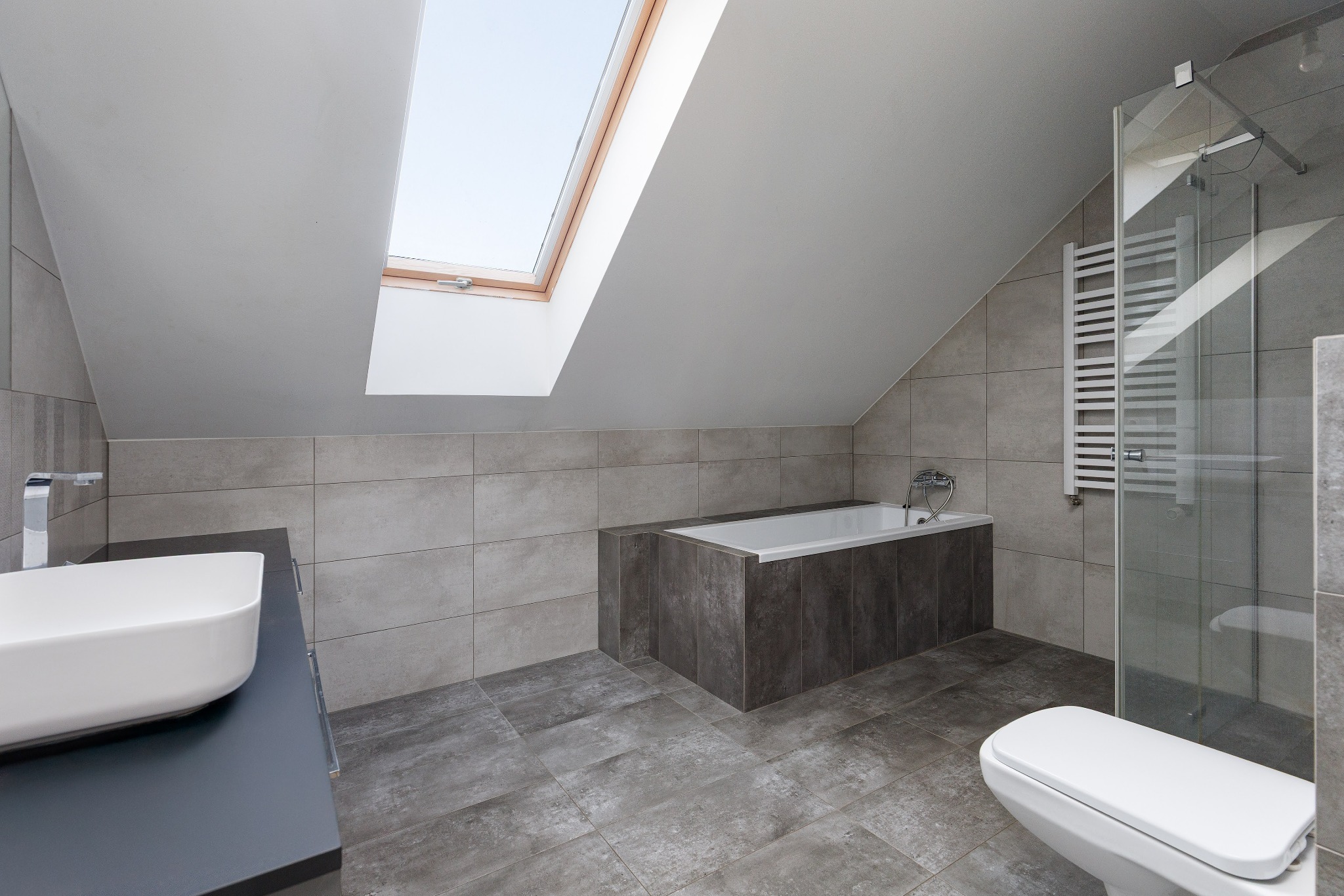 Nowoczesna łazienka na poddaszu z oknem dachowym, wykończona w szarościach. Umywalka nablatowa, wanna obudowana płytkami, kabina prysznicowa ze szklaną ścianką.