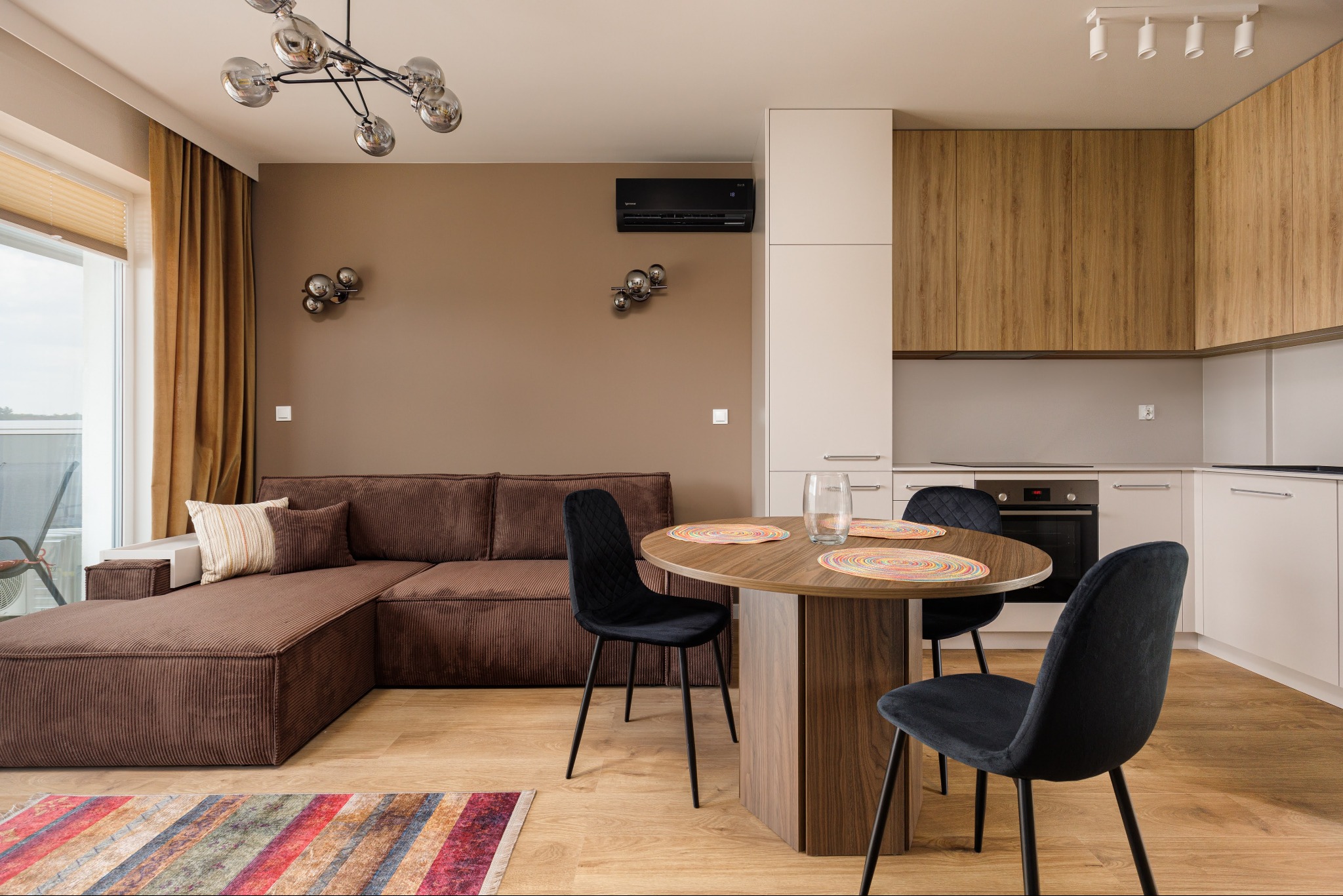 Jasne, przestronne wnętrze mieszkania z brązową sofą, okrągłym stołem i nowoczesną kuchnią w odcieniach bieli i drewna. Widok na balkon i designerskie oświetlenie.