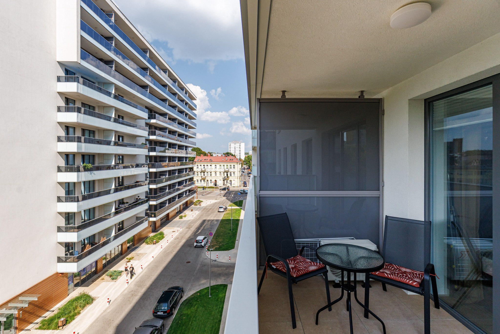 Nowoczesny balkon z widokiem na miasto. Szklany stolik i krzesła z poduszkami. W tle budynek apartamentowy i ulica z samochodami. Roleta przeciwsłoneczna częściowo zasłonięta.