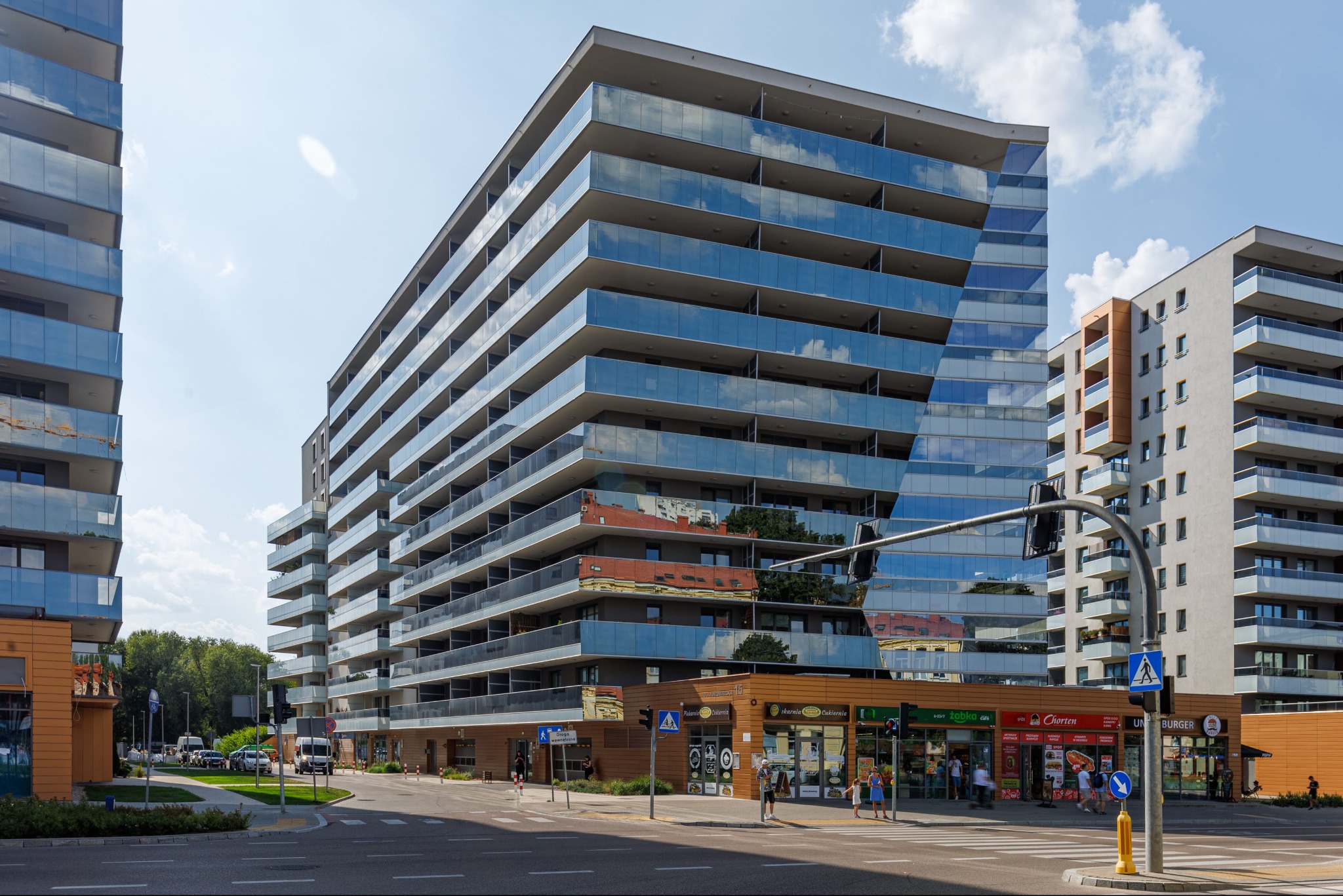 Nowoczesny budynek apartamentowy z balkonami i szklaną fasadą odbijającą niebo, widok z poziomu ulicy, z przejściem dla pieszych i sklepami w parterze.