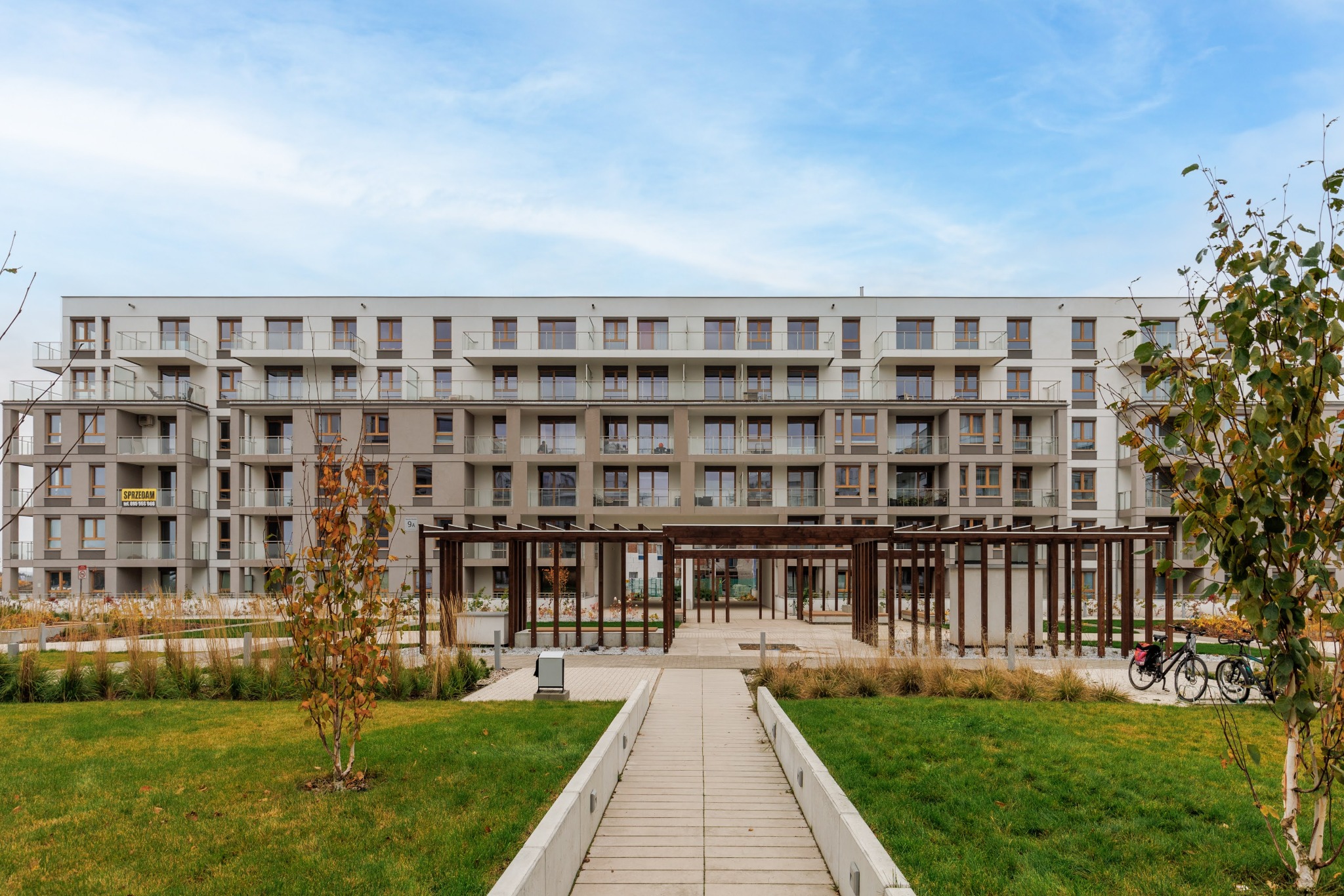 Nowoczesny apartamentowiec z balkonami, widok z parku z alejką i pergolą. Na balkonie widoczna tabliczka 'Sprzedam'.
