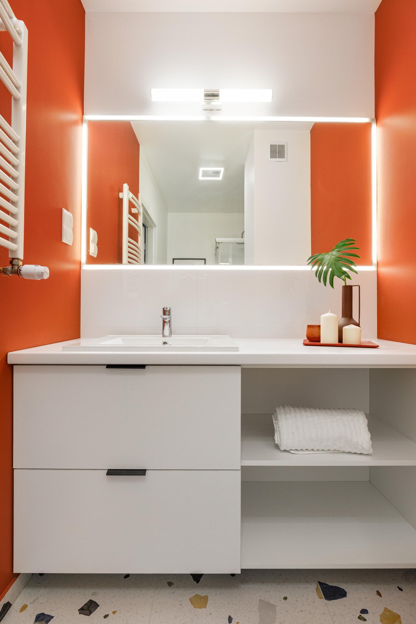 Nowoczesna łazienka z pomarańczowymi ścianami, białymi meblami i lustrem z podświetleniem LED. Na blacie dekoracje: wazon z liściem, świece i ręcznik na półce.
