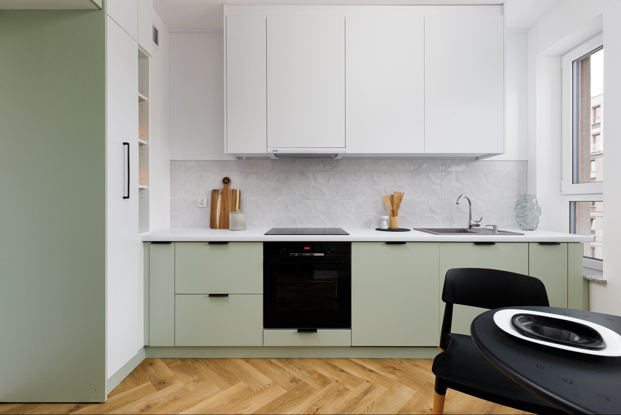 Nowoczesna kuchnia w odcieniach bieli i zieleni z drewnianą podłogą w jodełkę. Minimalistyczny design, widok z perspektywy stołu w Białymstoku. Jasne wnętrze.