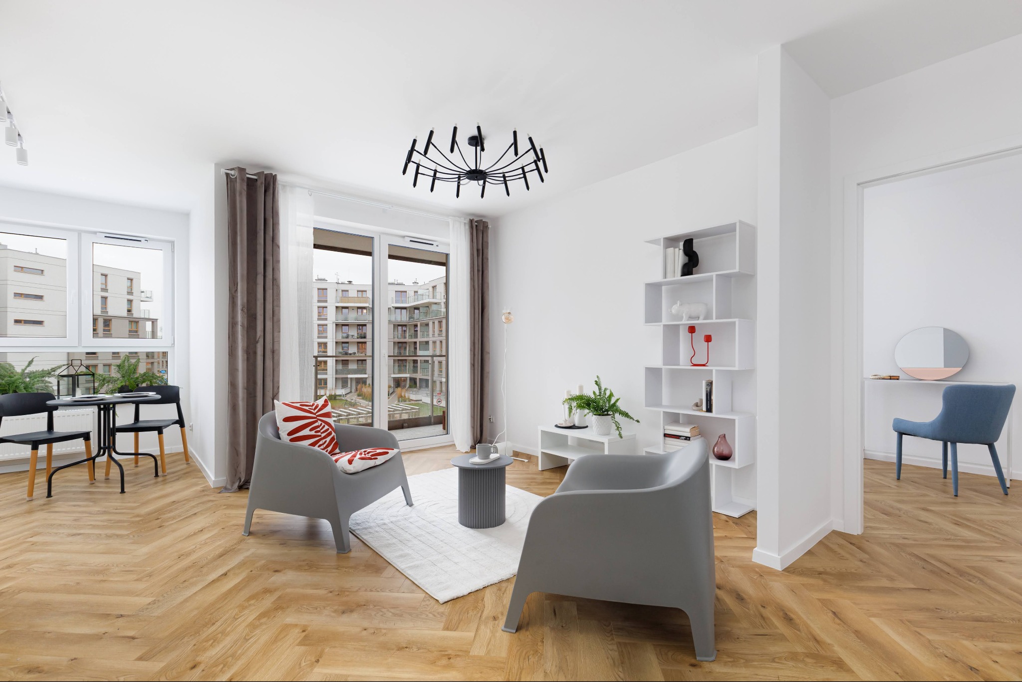 Jasne wnętrze mieszkania z parkietem w jodełkę, szarymi fotelami i widokiem na osiedle przez okno. Minimalistyczny design z akcentami roślinnymi i nowoczesnym oświetleniem.