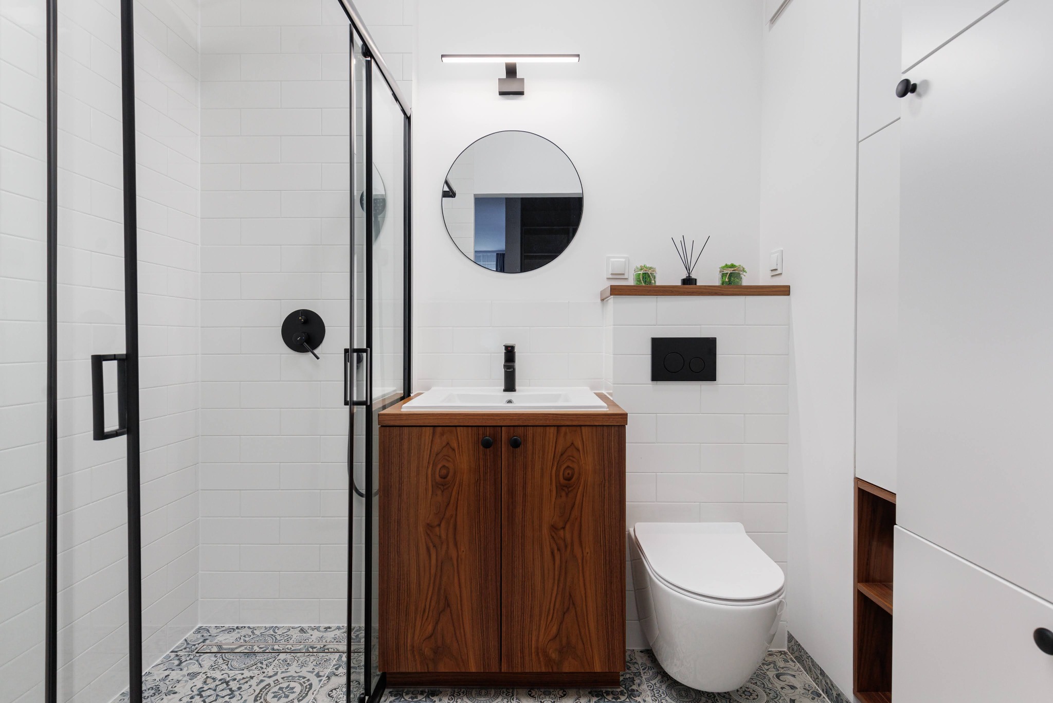 Nowoczesna łazienka z kabiną prysznicową typu walk-in, umywalką z szafką z drewna orzechowego i okrągłym lustrem. Białe płytki i wzorzysta podłoga.