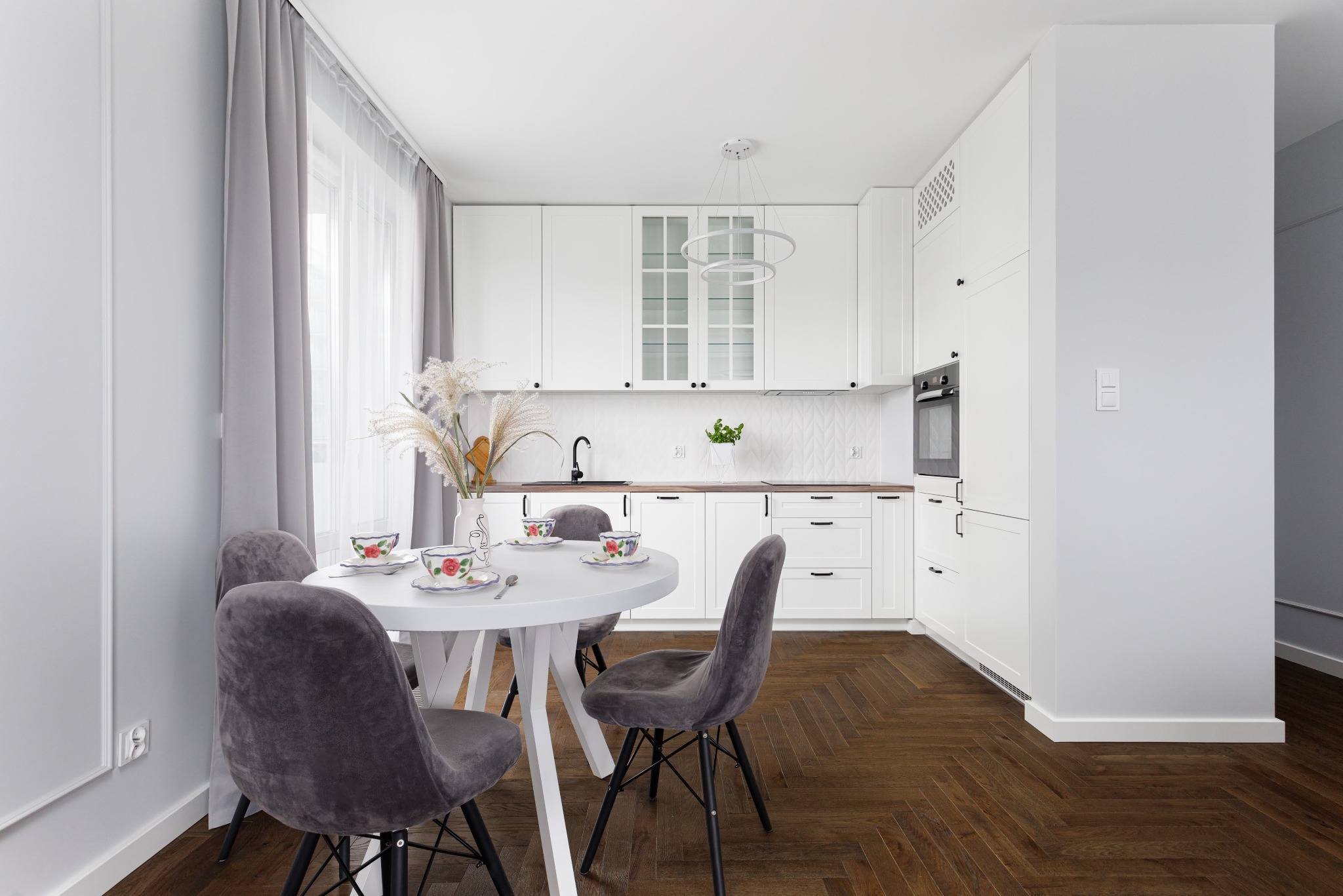 Jasna kuchnia z białymi meblami i drewnianym blatem, okrągły stół z szarymi krzesłami i zastawą. Parkiet w jodełkę. Minimalistyczny design, nowoczesne oświetlenie.