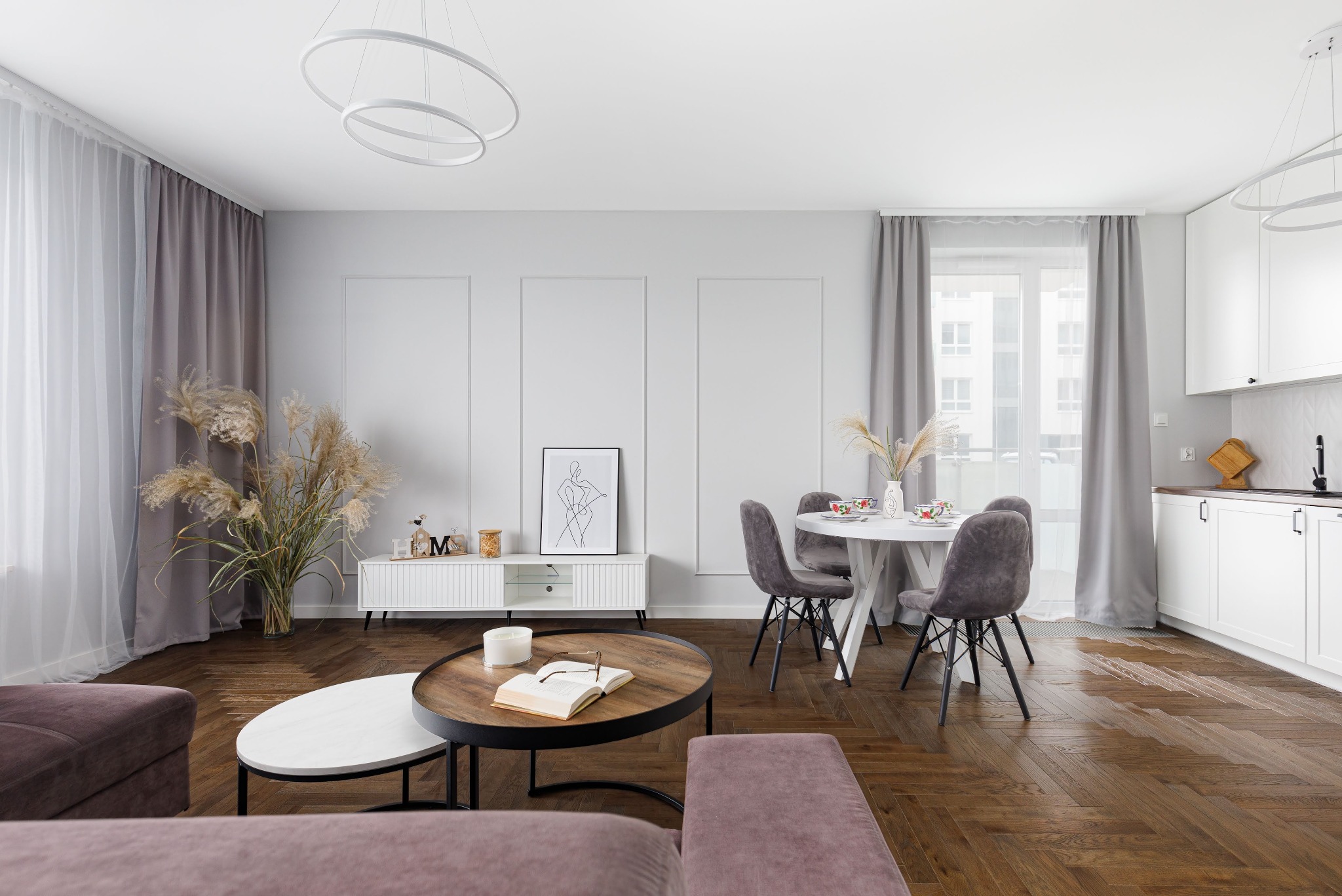 Jasne wnętrze mieszkania z parkietem w jodełkę. Okrągły stół, krzesła, sofa i stoliki kawowe. Dekoracje: trawa pampasowa, grafika na ścianie, książka na stoliku.