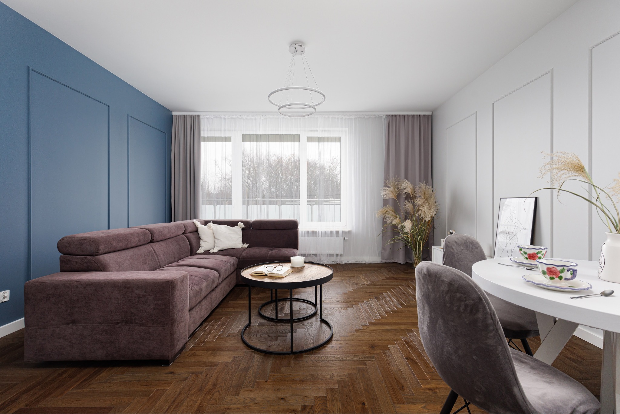 Jasne wnętrze: brązowa sofa narożna na parkiecie w jodełkę, okrągły stolik kawowy, biały stół z nakryciem, niebieska ściana z dekoracyjnymi listwami.
