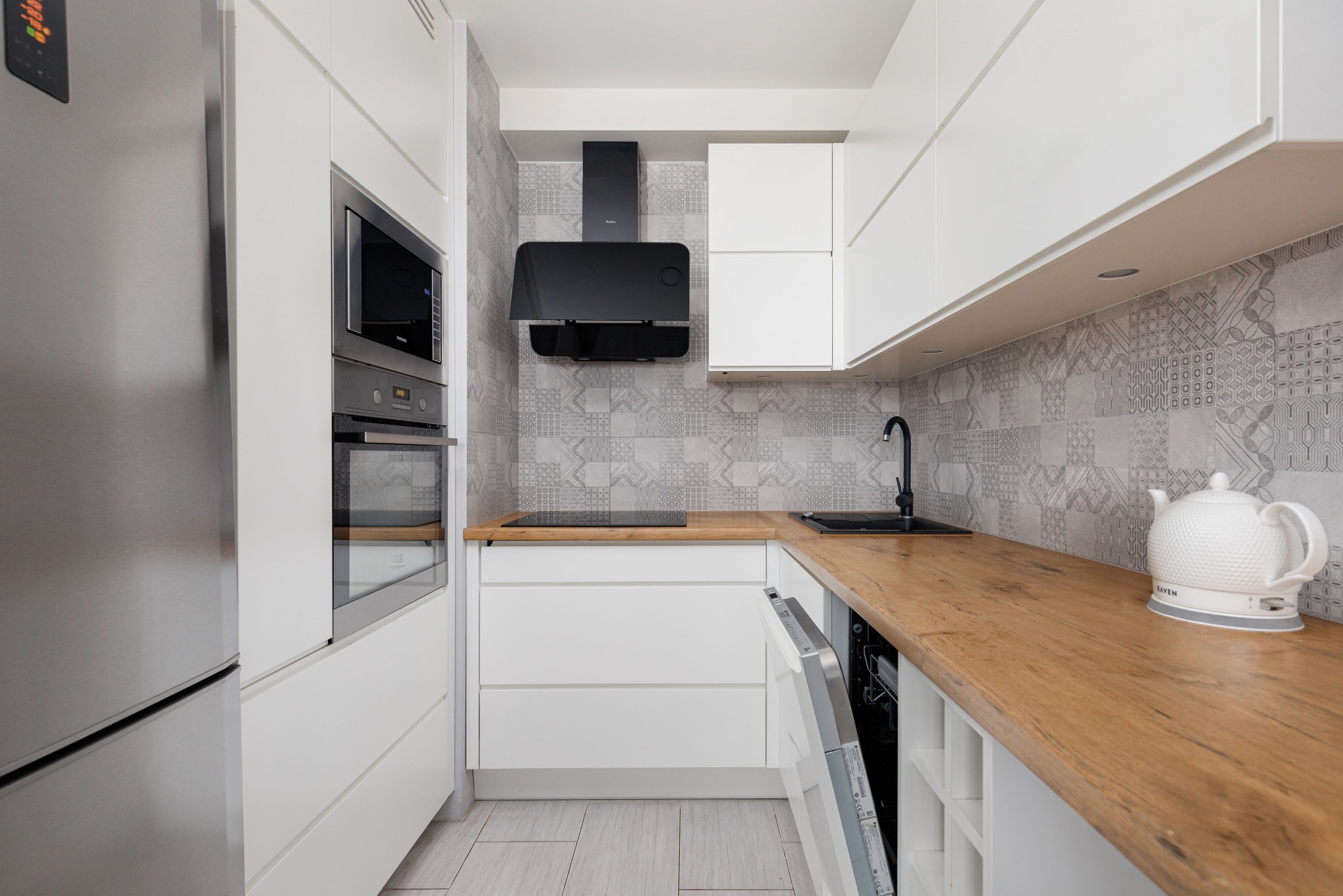 Nowoczesna kuchnia w bieli i drewnie: zabudowa z piekarnikiem, mikrofalówką, płytą indukcyjną i okapem. Na blacie czajnik elektryczny. Otwarta zmywarka.