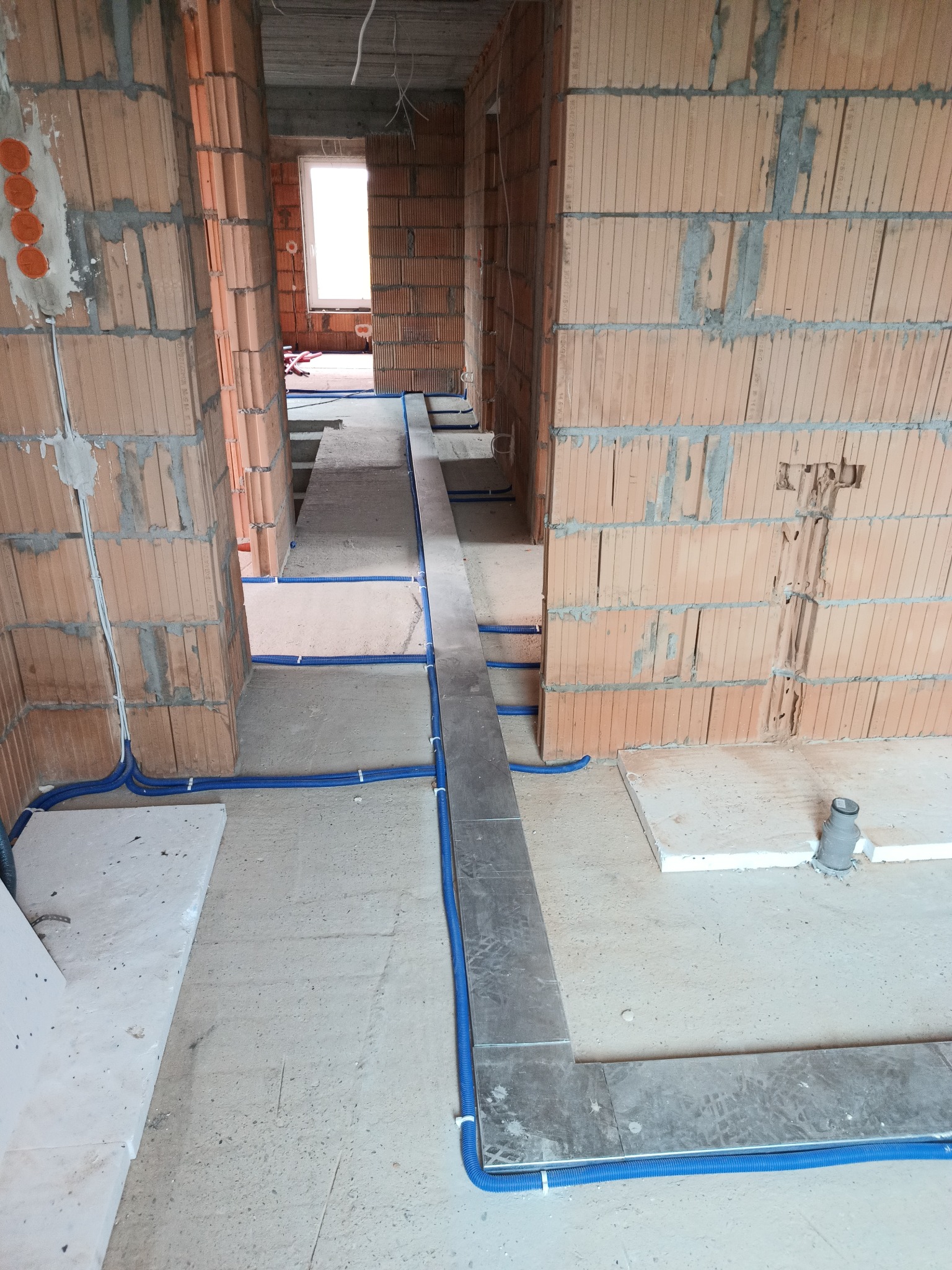 Niebieskie rury instalacyjne prowadzone wzdłuż surowych ścian z cegły i podłogi w trakcie budowy. Widoczne kable i puszki elektryczne w ścianach.