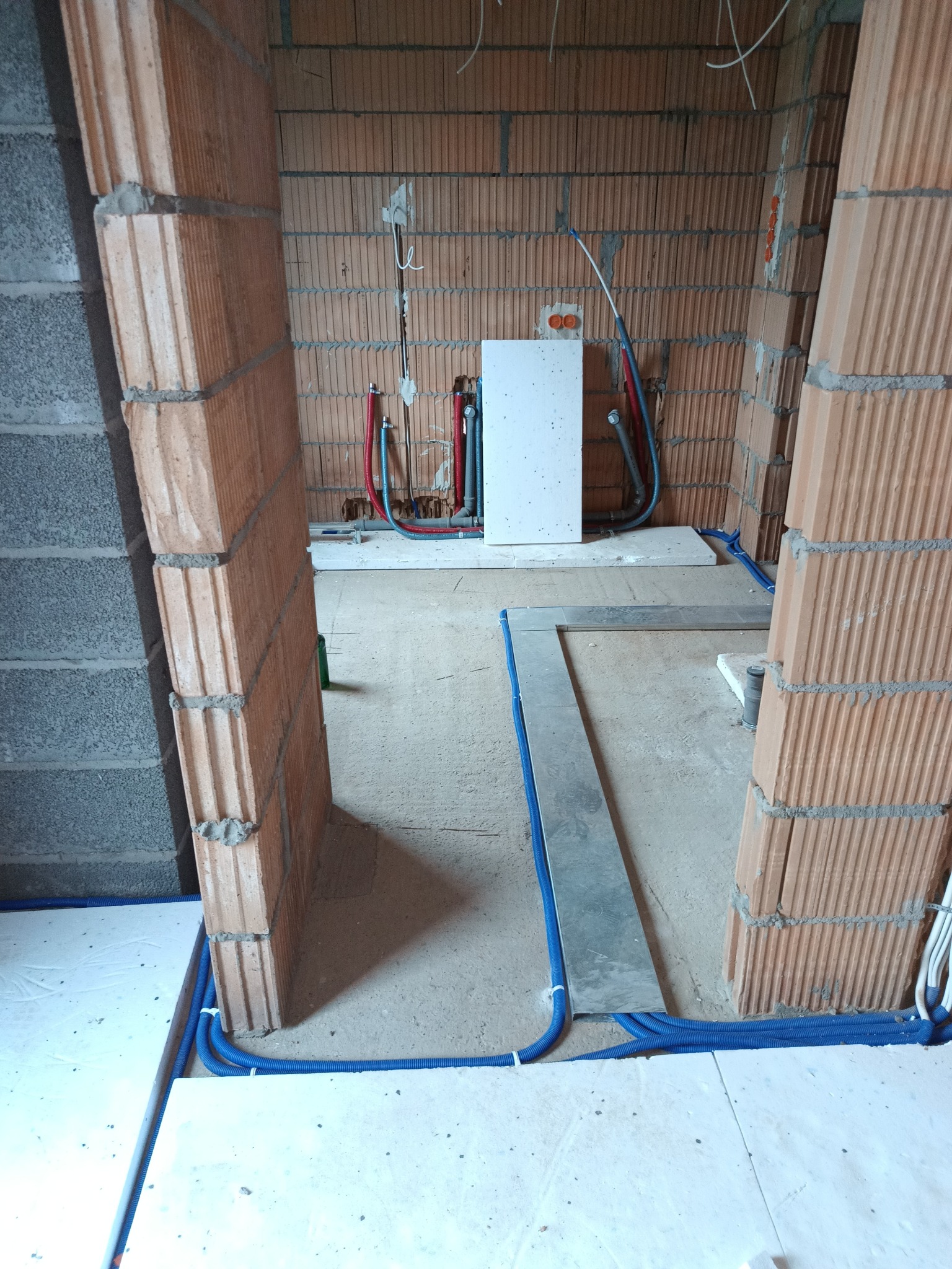 Wnętrze w trakcie prac instalacyjnych: widoczne rury w peszlach na podłodze, instalacja wodna i elektryczna na ścianie z cegły dziurawki. Surowy stan.