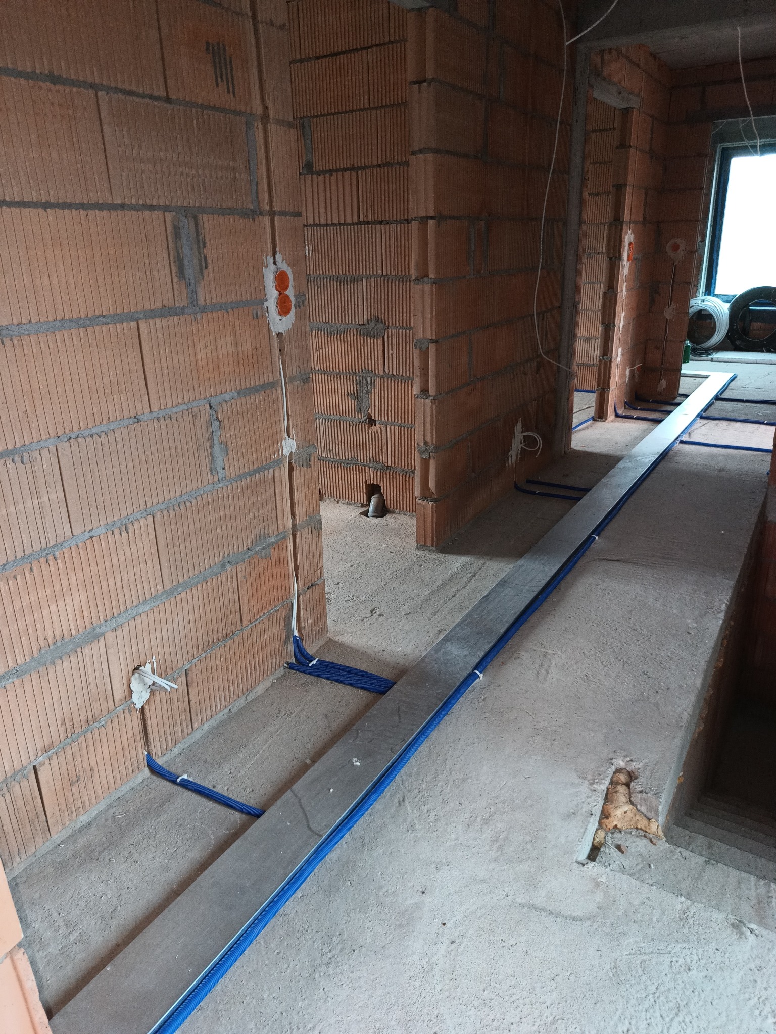 Wnętrze w trakcie budowy: widoczne ceglane ściany, bruzdy z niebieskimi rurami instalacyjnymi i puszki elektryczne w ścianach. Surowy beton na podłodze.
