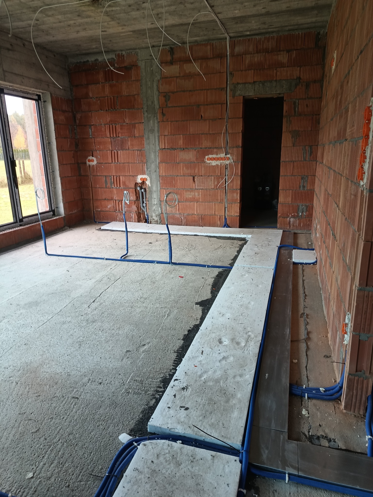 Surowy stan: Instalacja elektryczna w peszlach na podłodze i ścianach w nowym budynku z cegły, przed wylewką. Widoczne kable wystające z sufitu i ściany.