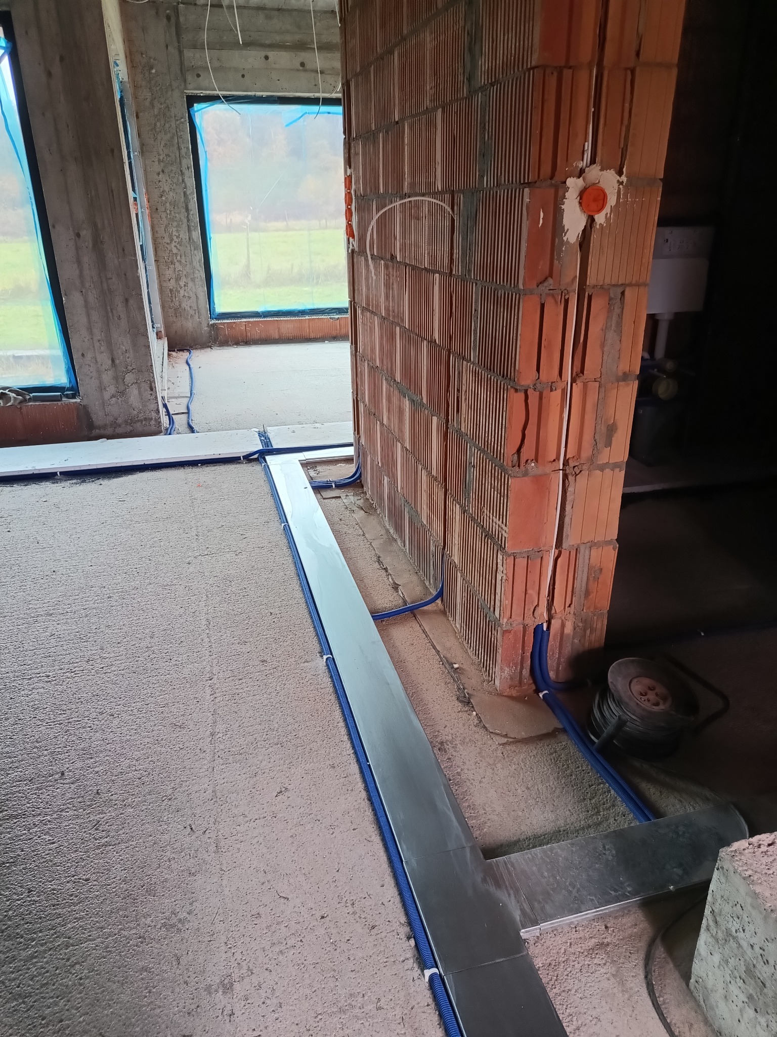 Niebieskie rury w peszlach ułożone wzdłuż ściany z czerwonej cegły dziurawki na surowym betonie w trakcie budowy. Widoczne okna z niebieską folią.