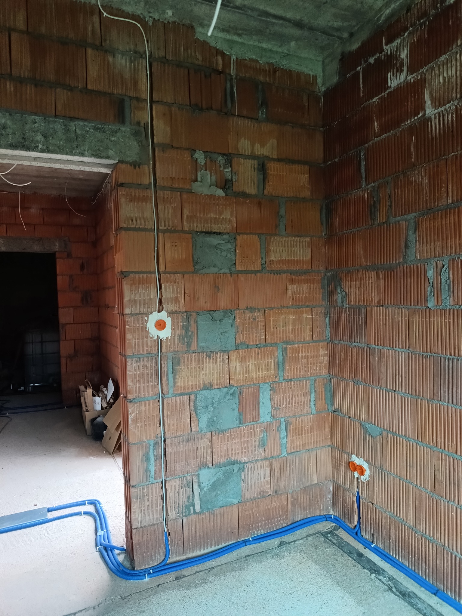 Surowa ściana z cegły z widoczną instalacją elektryczną: kable w peszlach i puszki pod gniazdka. Niebieskie rury peszlowe na podłodze. W tle widoczne wejście.