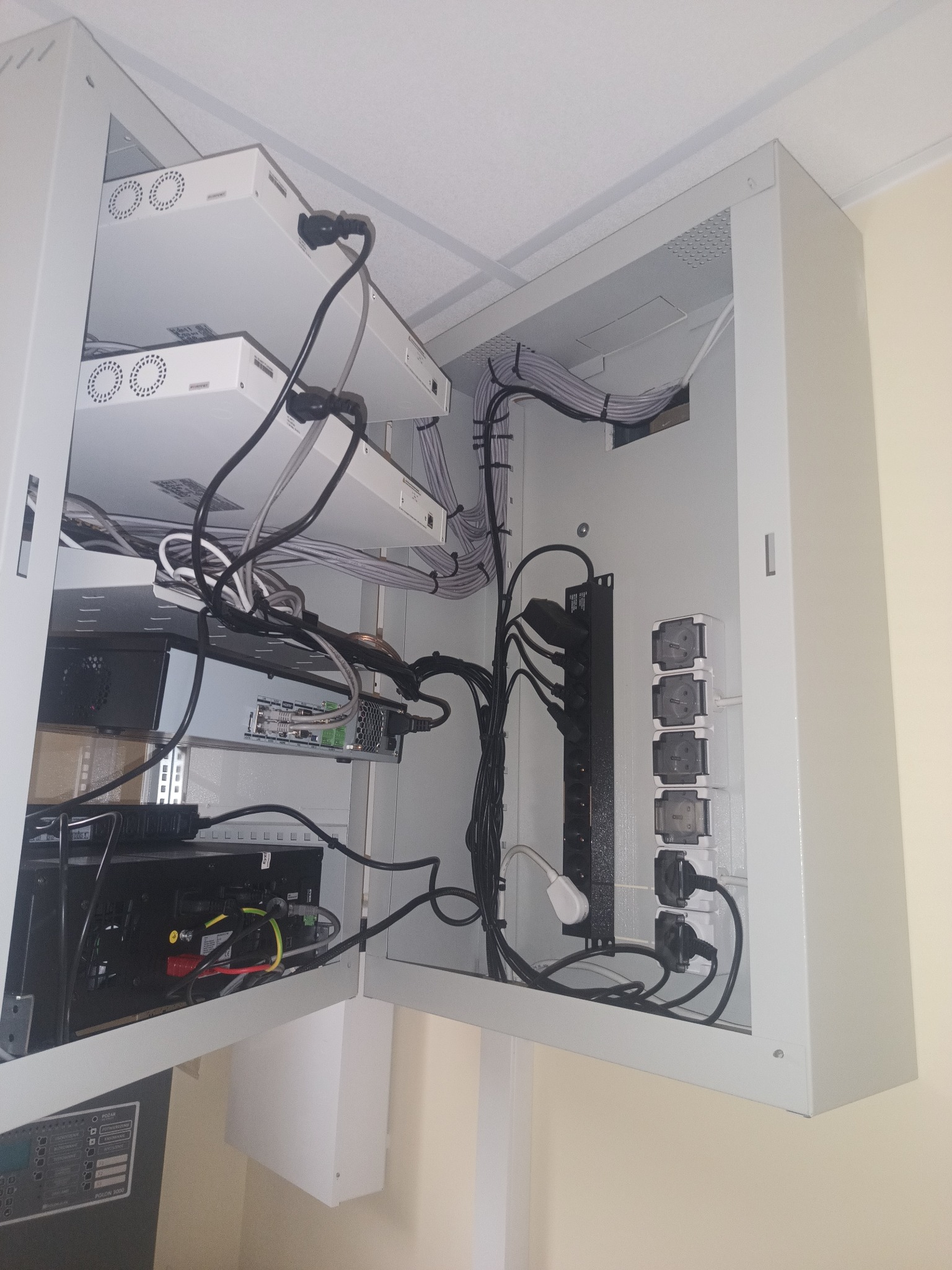Otwarta szafa rack z zamontowanymi urządzeniami sieciowymi, listwą zasilającą i uporządkowanym okablowaniem. Widoczne gniazda elektryczne i elementy systemu alarmowego.