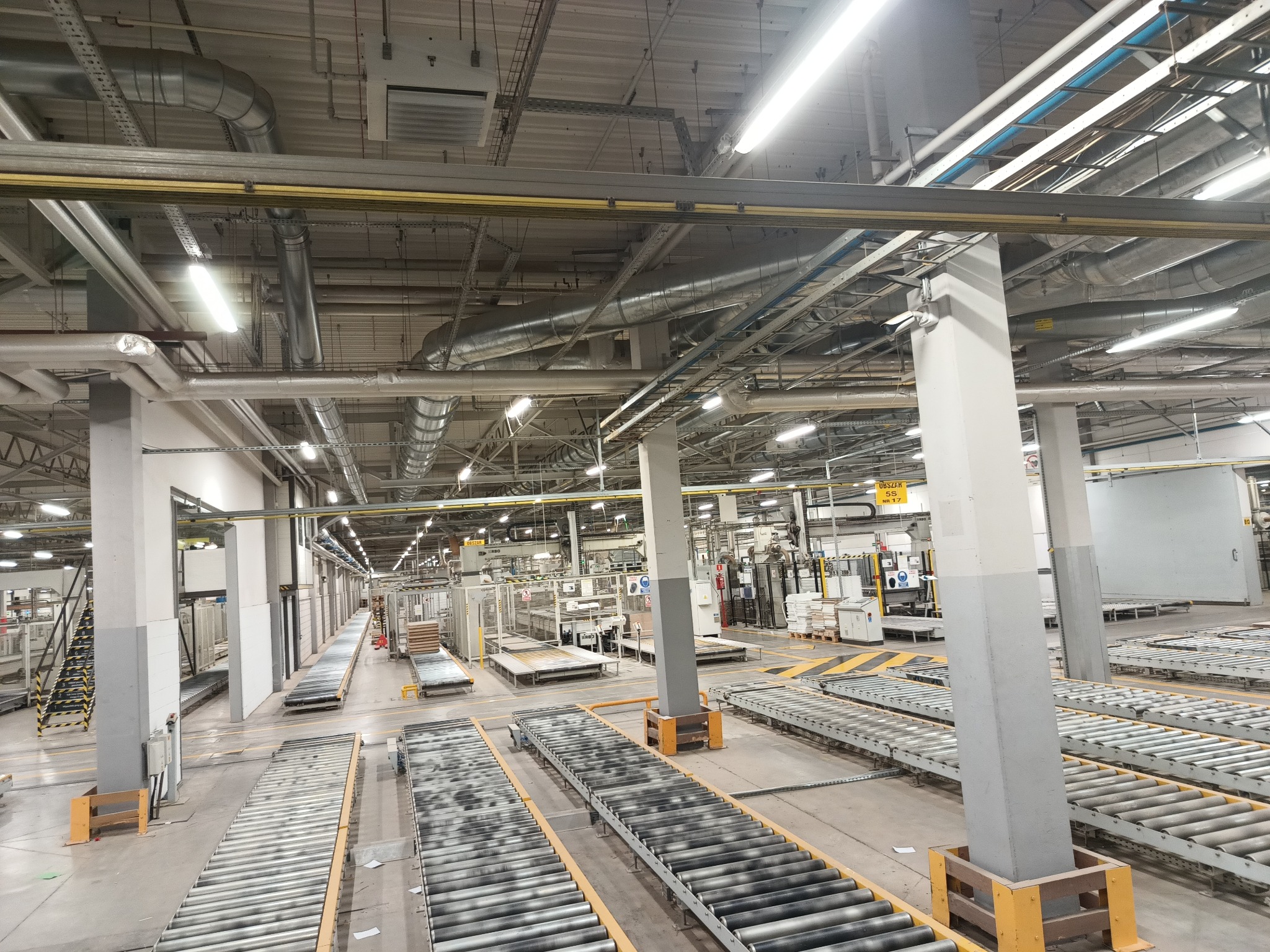 Rozległa hala fabryczna z systemem przenośników rolkowych, oświetlona lampami jarzeniowymi i z widocznymi instalacjami pod sufitem. Perspektywa z góry.