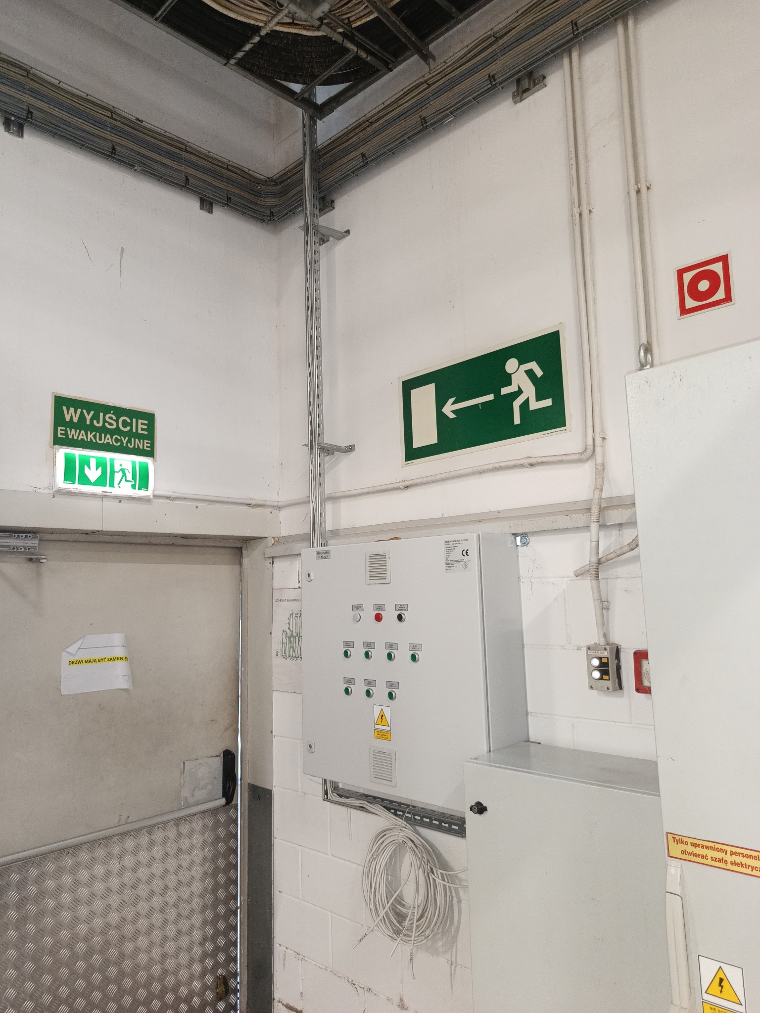 Szafa sterownicza z okablowaniem, oznaczenia wyjścia ewakuacyjnego i przeciwpożarowe w pomieszczeniu technicznym. Widoczne kable w korytkach i rurach na ścianach.