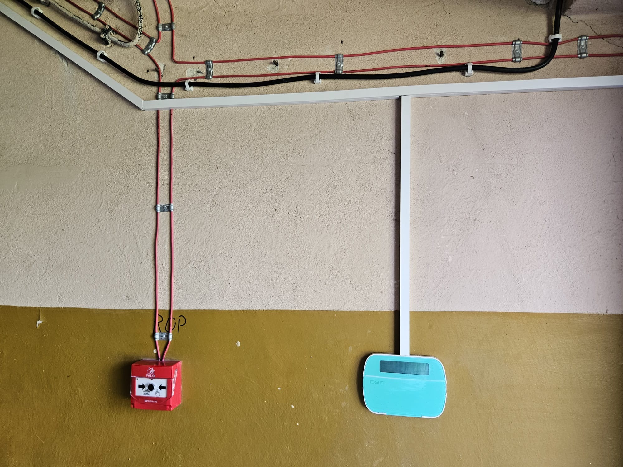 Czerwony ręczny ostrzegacz pożarowy ROP i panel alarmowy DSC na ścianie z instalacją elektryczną. Widoczne kable w peszlach i uchwytach.