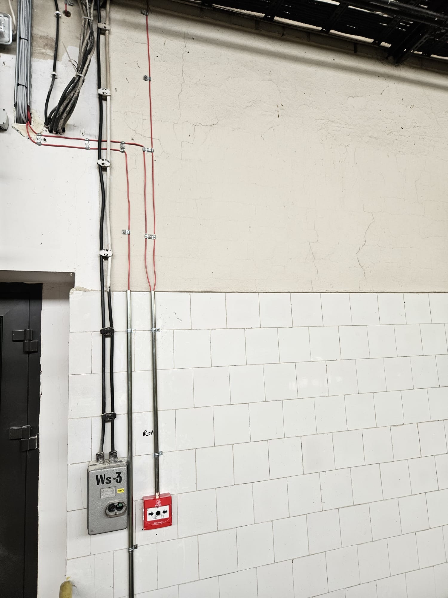 Szczegółowe ujęcie instalacji elektrycznej z przewodami w peszlach i puszką przyłączeniową na ścianie wyłożonej białymi płytkami, z widocznym czujnikiem pożarowym.