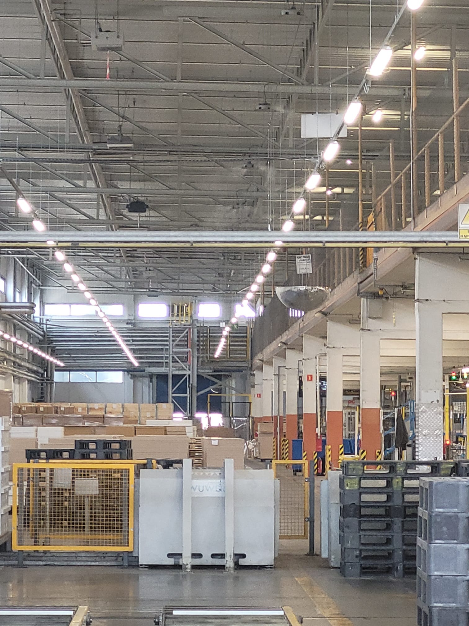Oświetlenie hali produkcyjnej w Pilchowicach: rzędy lamp wiszących pod sufitem, widoczne elementy konstrukcyjne i okna w tle. Perspektywa z poziomu podłogi.