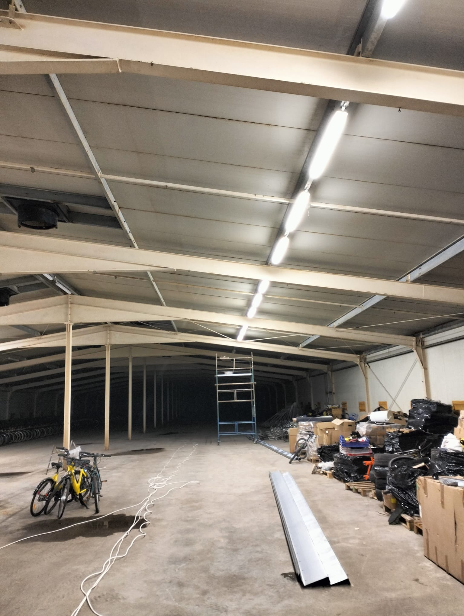 Wnętrze hali z widoczną konstrukcją dachu, oświetleniem, rowerami i elementami instalacji elektrycznej. Widoczna drabina i kable. Hala w trakcie prac instalacyjnych.