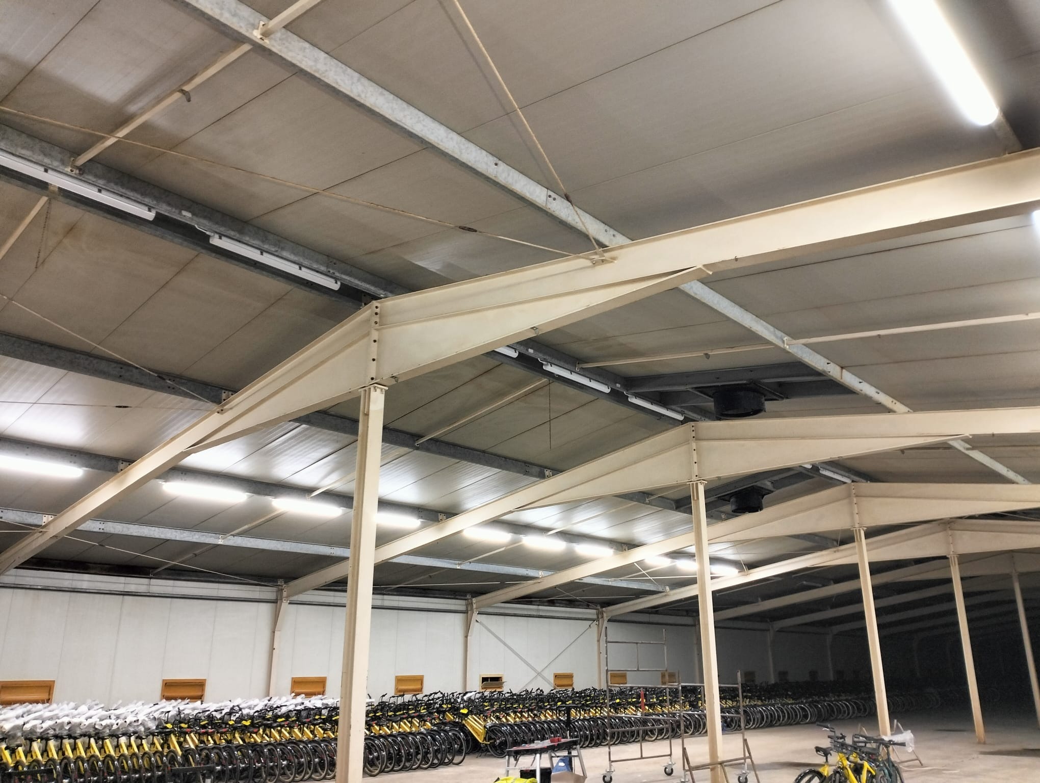 Wnętrze hali magazynowej z oświetleniem LED pod sufitem. Widoczne metalowe konstrukcje wsporcze i rzędy rowerów w tle. Jasne, równomierne światło.