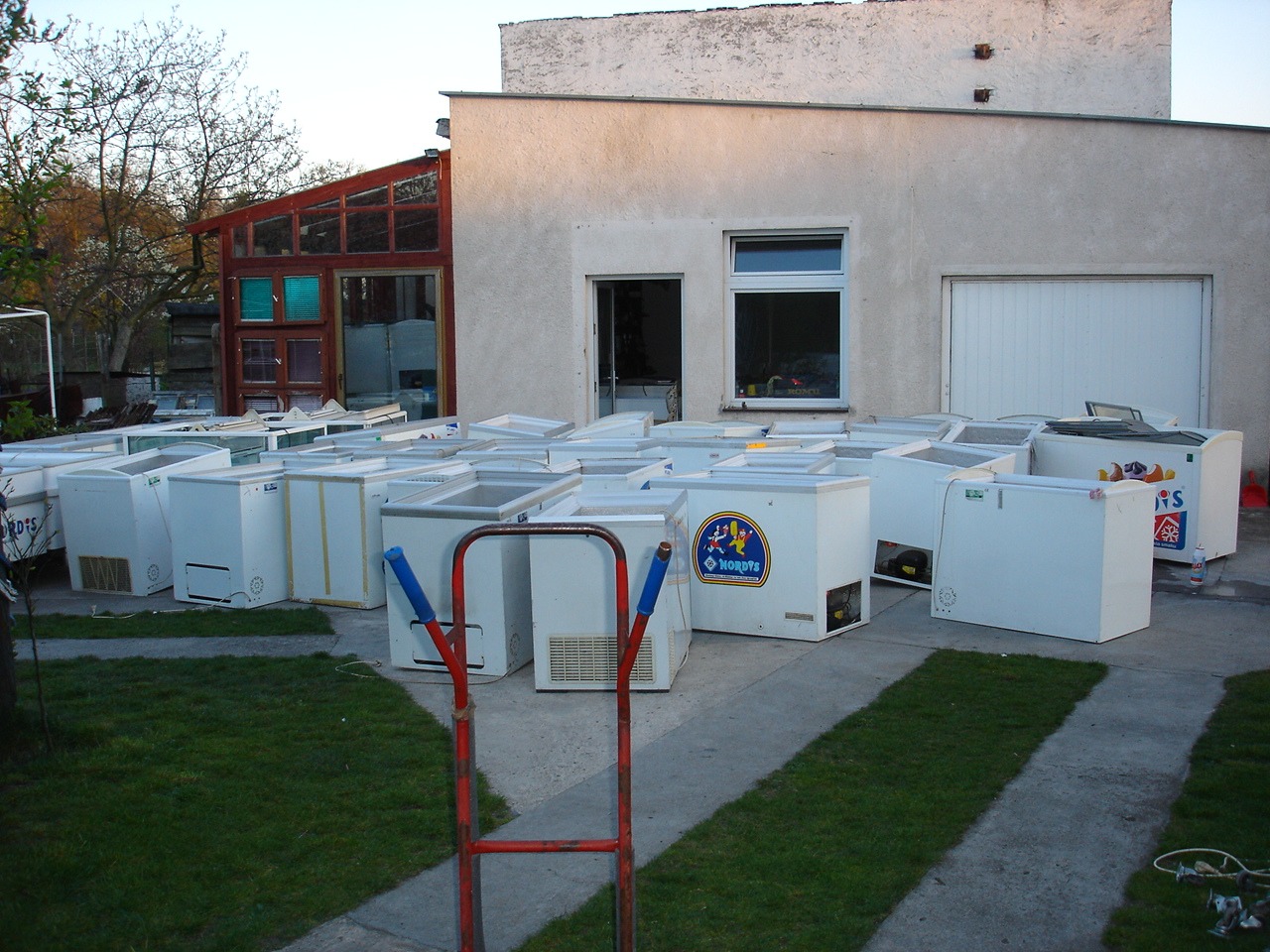 Magazyn białych, używanych zamrażarek skrzyniowych na zewnątrz budynku. W tle widoczny budynek gospodarczy z oknem i wrotami garażowymi.