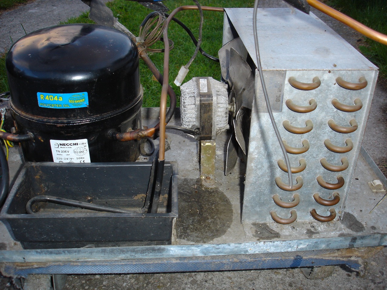 Otwarty agregat chłodniczy z czarnym kompresorem, widoczną tabliczką znamionową i chłodnicą z wentylatorem. Widoczne elementy instalacji i okablowanie.