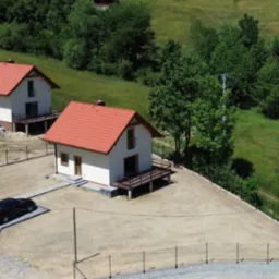 Widok z góry na osiedle domów jednorodzinnych z czerwonymi dachami, otoczonych ogrodzeniem i zielenią. Jeden z domów ma zaparkowany samochód.