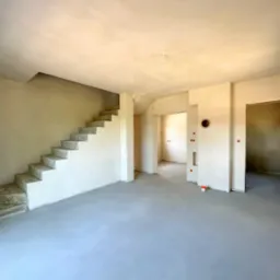 Surowe wnętrze domu w trakcie wykańczania: schody betonowe, wylewka na podłodze, ściany w tynku, widoczne otwory drzwiowe i przejścia. Minimalistyczny, surowy styl.