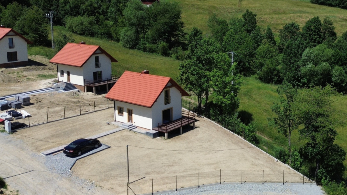 Widok z góry na osiedle domów jednorodzinnych z czerwonymi dachami, otoczonych ogrodzeniem i zielenią. Jeden z domów ma zaparkowany samochód.